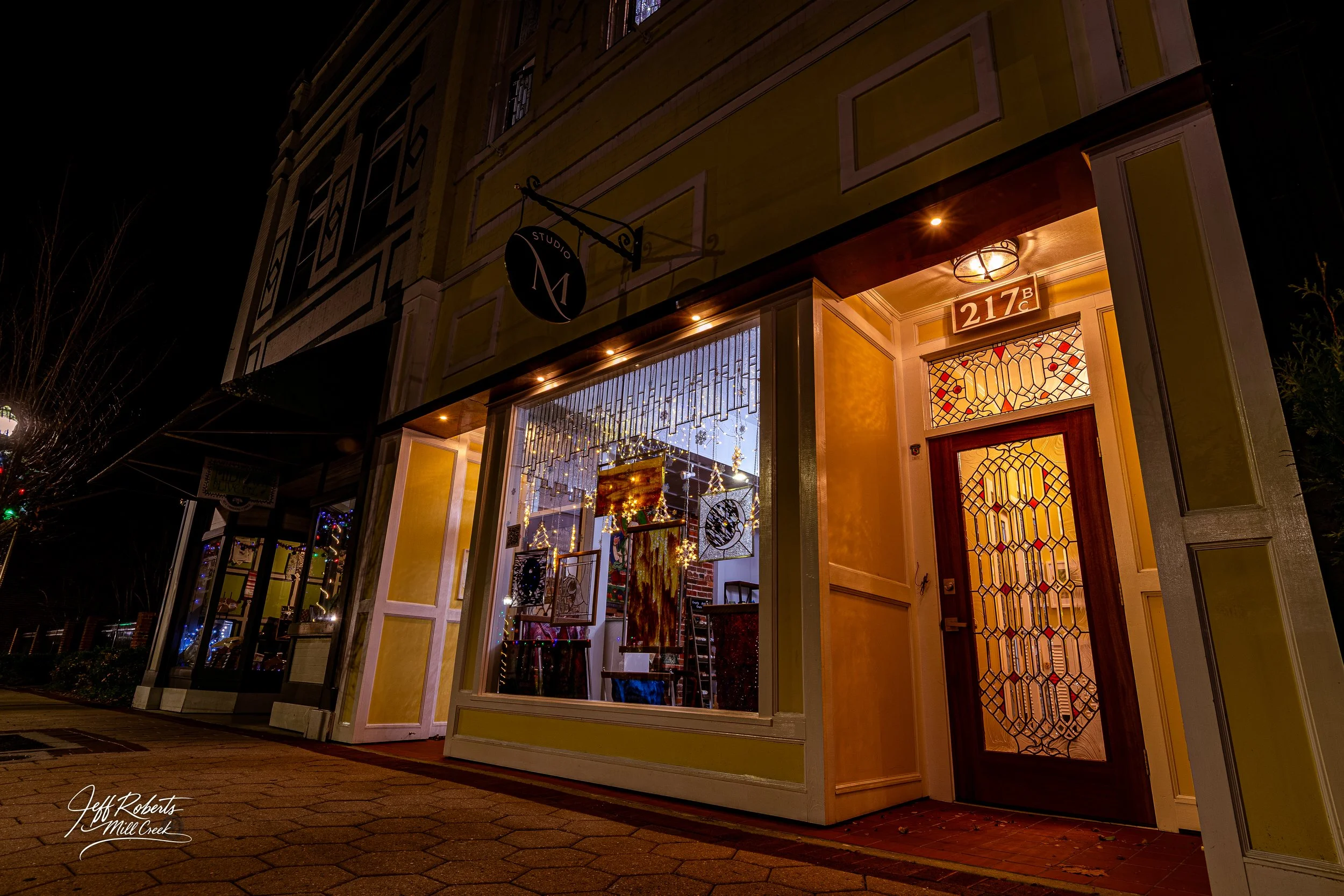 Storefront at night with large window display and ornate stained glass door, yellow exterior walls, decorative lighting, and sign reading 217B