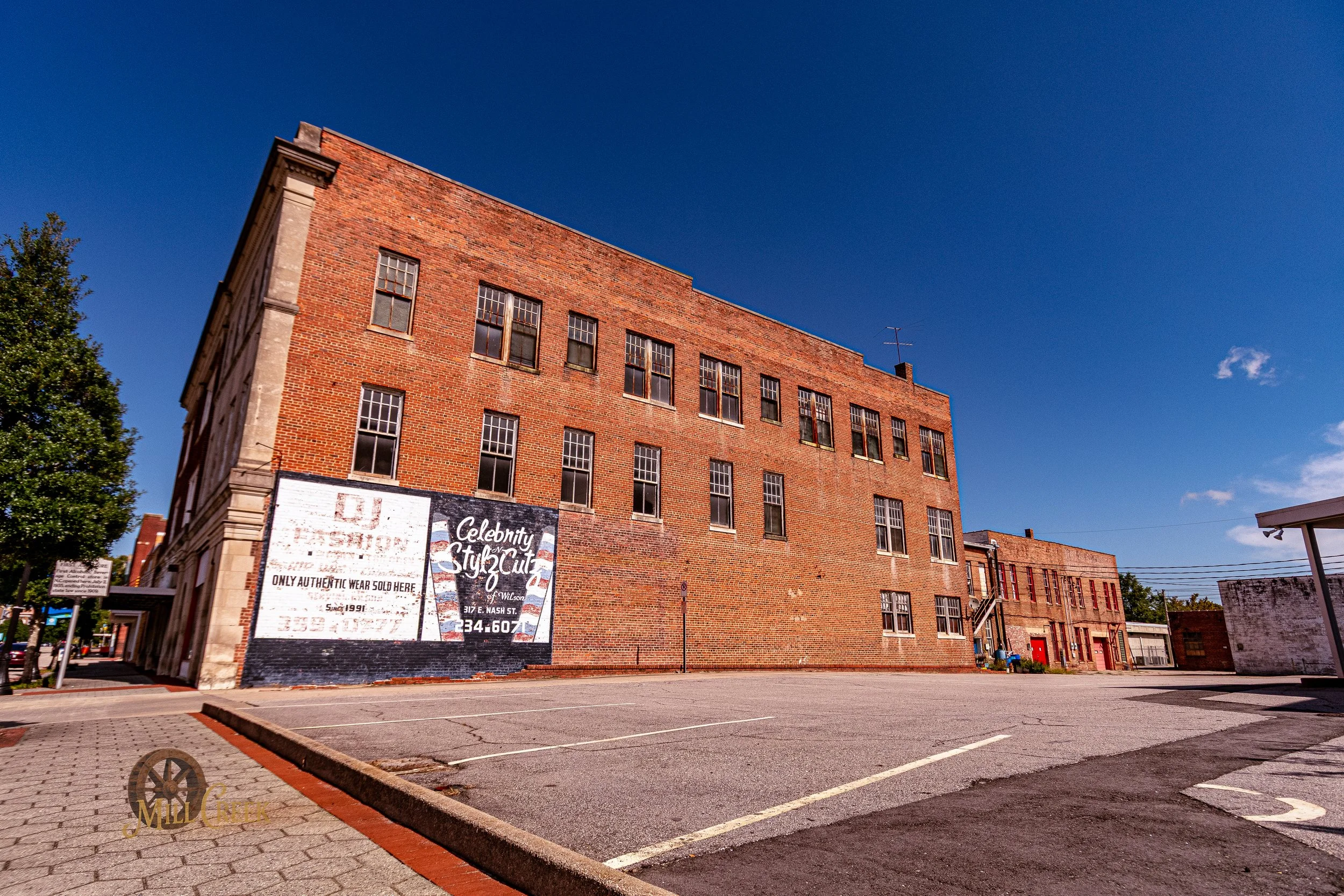 Empty parking lot in front of a large brick building with multiple windows and advertisements on the wall, under a clear blue sky.