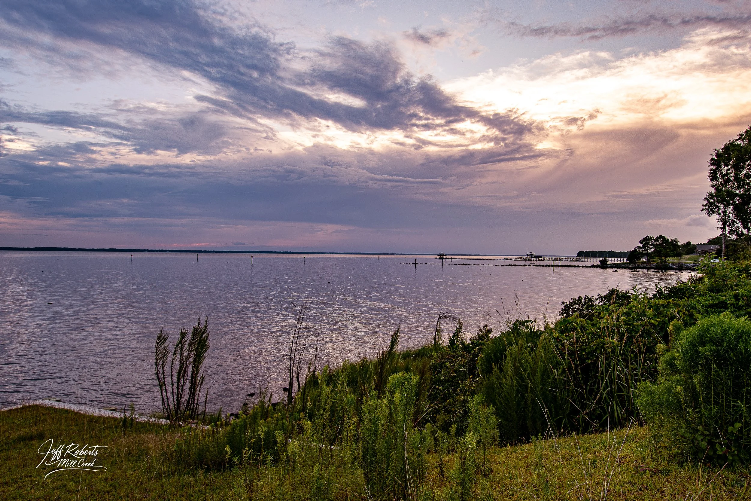Scenic view of a lake during sunset with cloudy sky, some greenery and plants in the foreground, a pier and small structures in the distance.