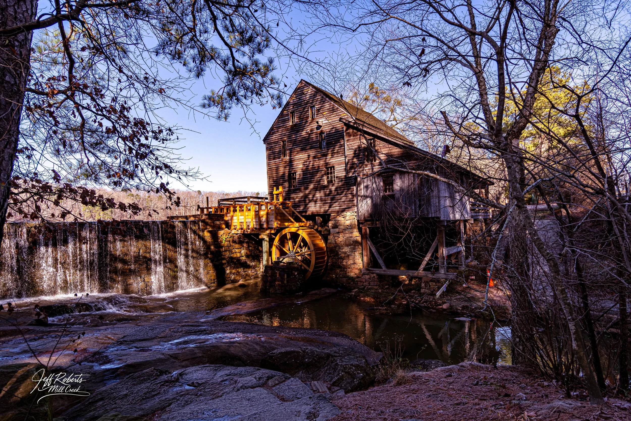 Old wooden watermill with a water wheel beside a small creek, surrounded by leafless trees in a rural setting.