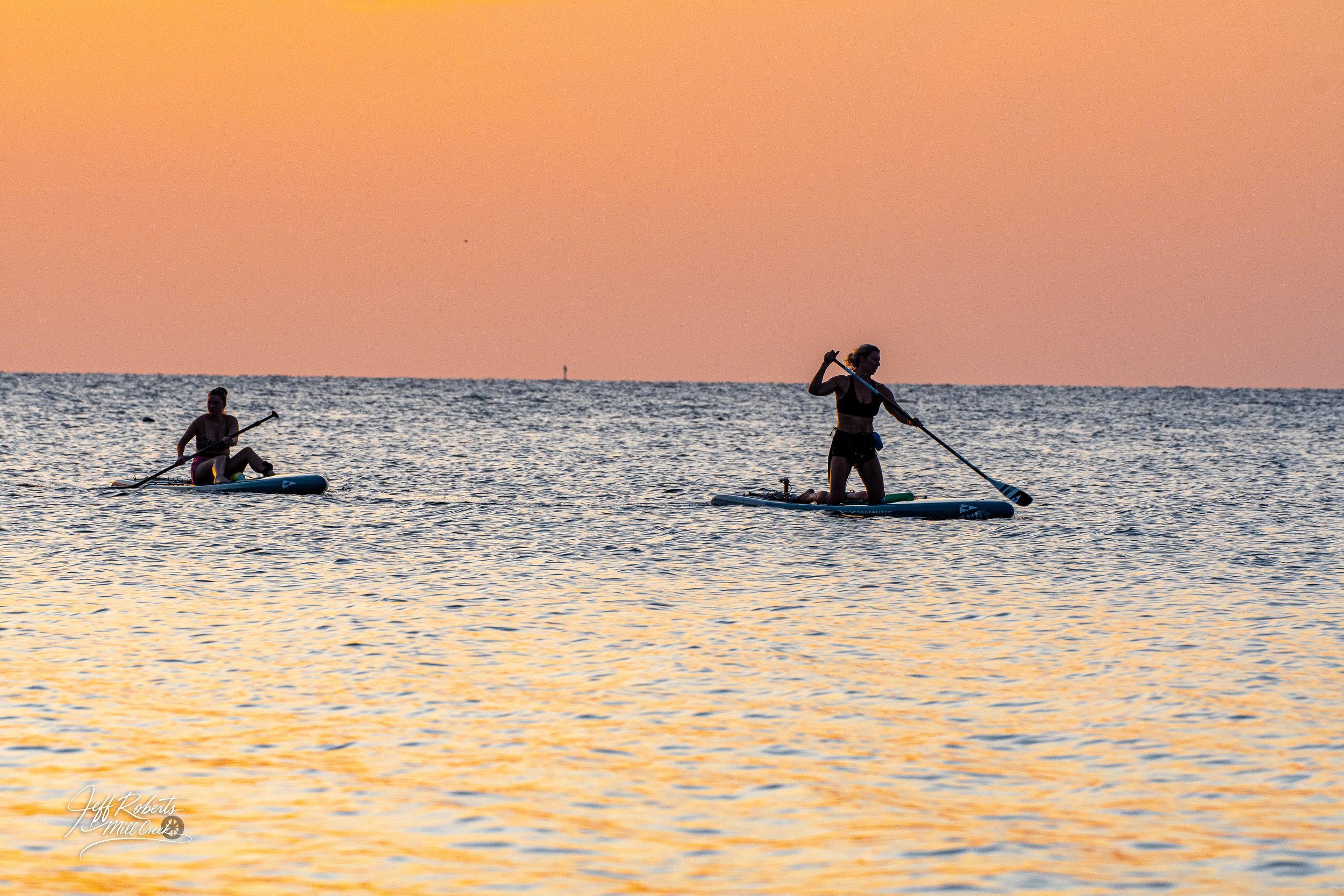 Two people paddleboarding on the water during sunset with a pastel sky in the background.