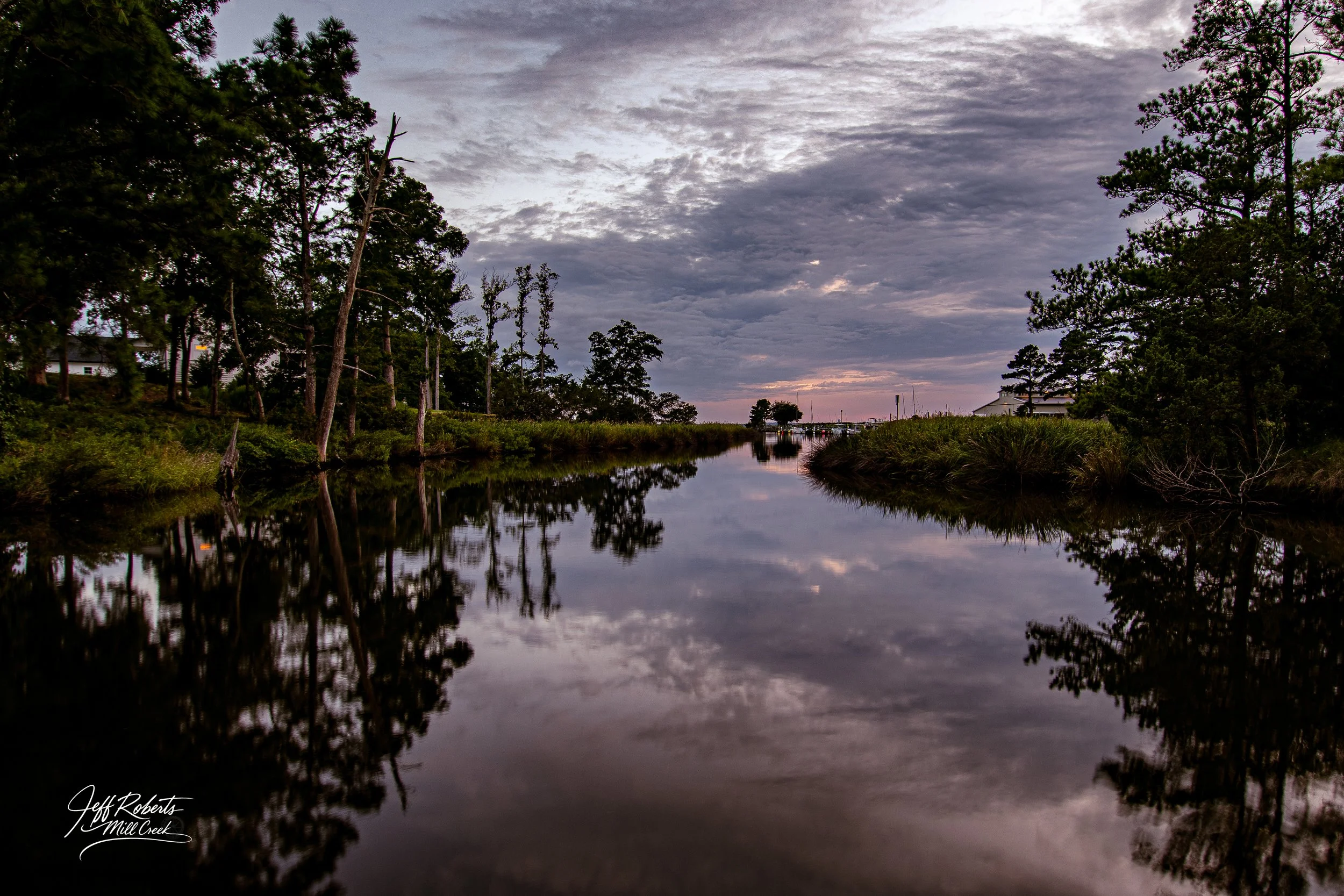 Calm waterway reflecting trees and cloudy sky during sunset, with houses and boats in the distance on Mill Creek in Jeff Roberts.