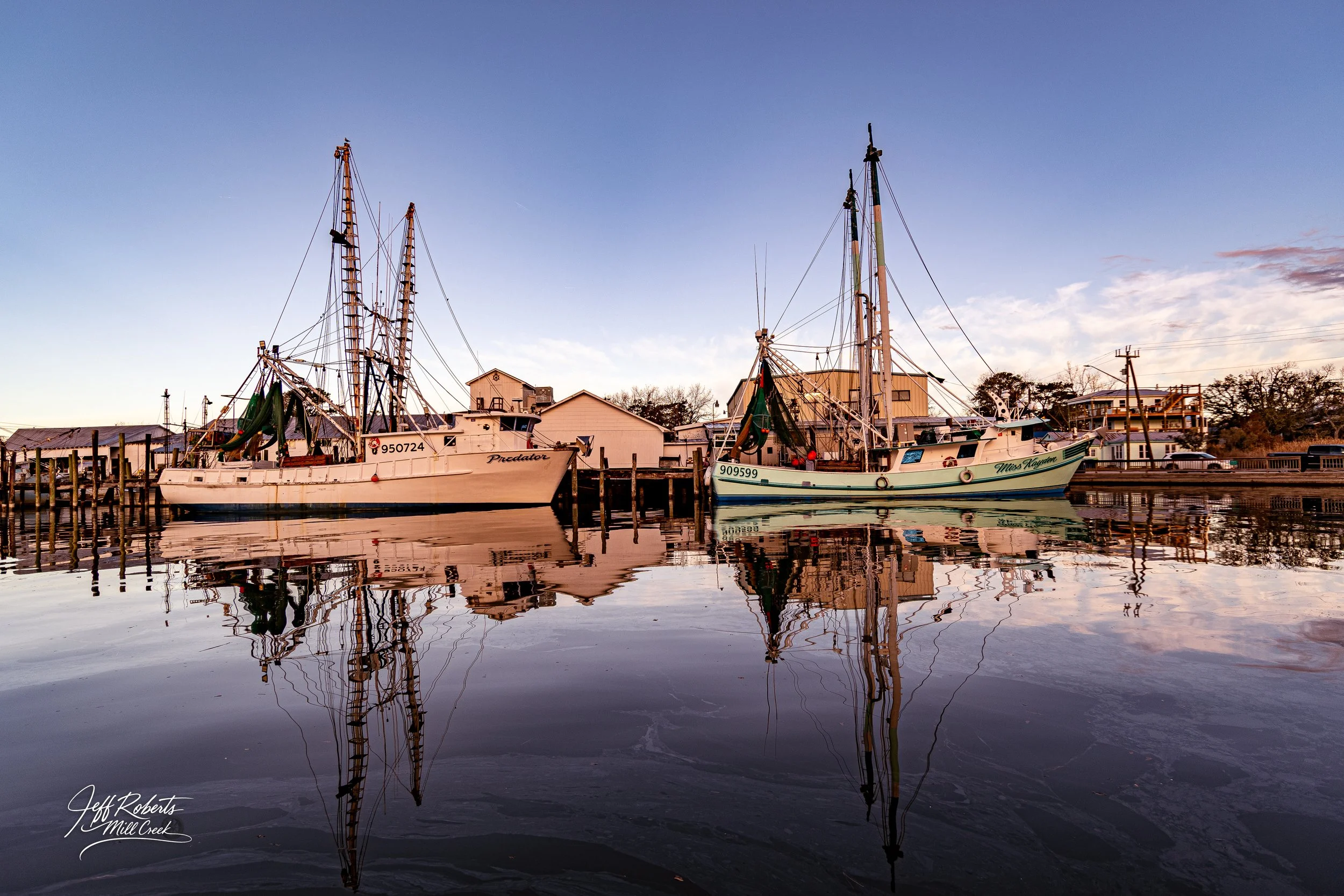 Two sailboats docked at a pier on calm water with reflections, houses in the background, and a clear sky.