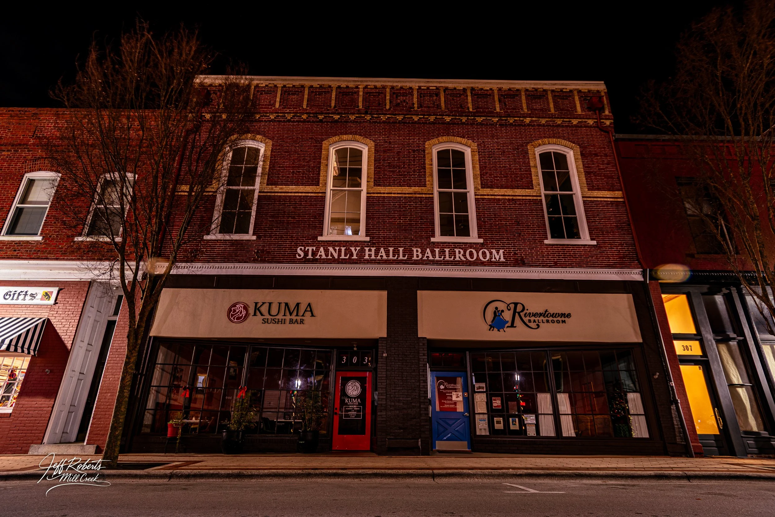 Night view of the brick building with a sign indicating Stanly Hall Ballroom; storefronts include Kuma Sushi Bar with a red door and Rivertowne Ballroom with a blue door, and a gift shop on the left.