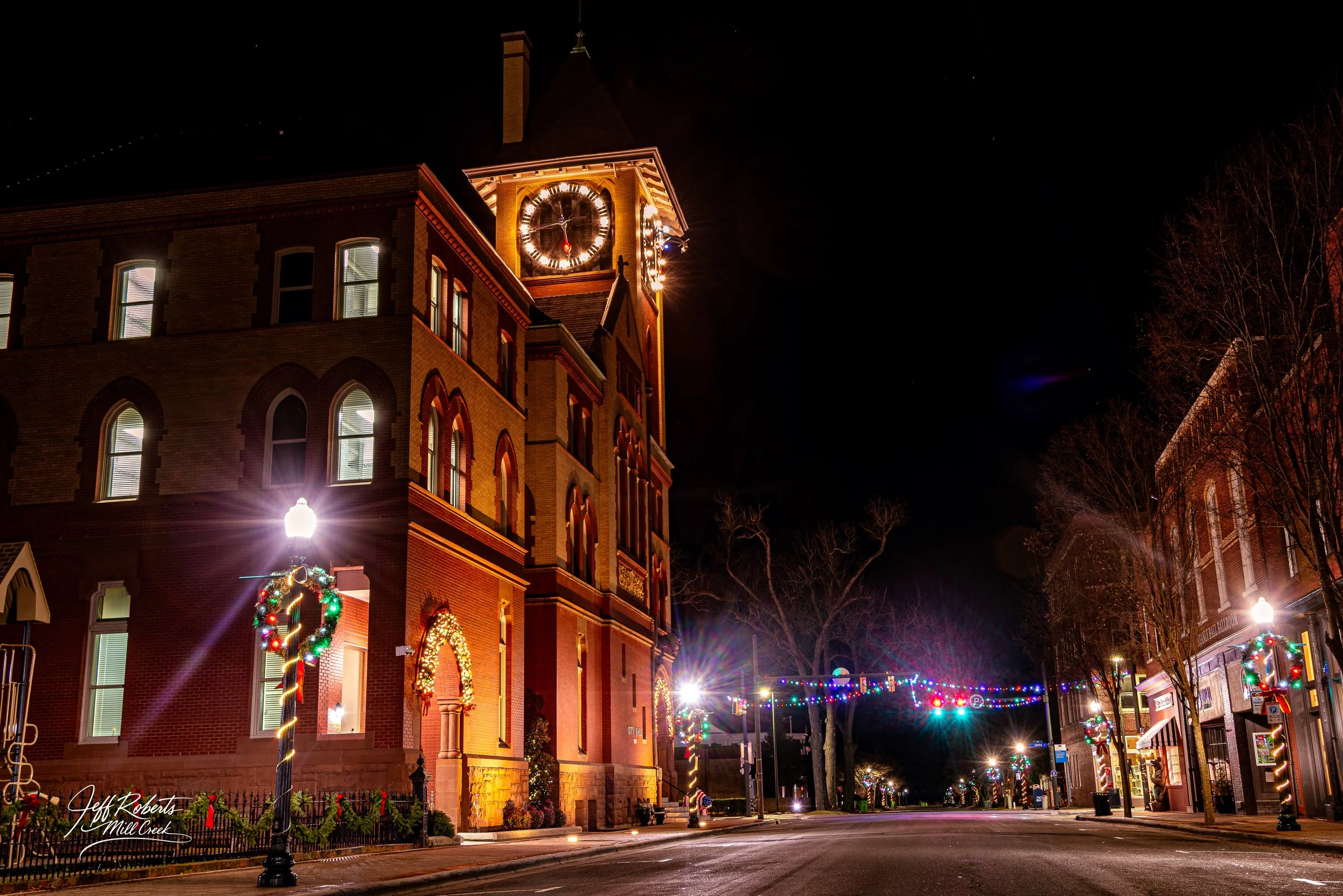 Nighttime street scene decorated with Christmas lights and wreaths, featuring a large brick building with a clock tower illuminated at the corner.
