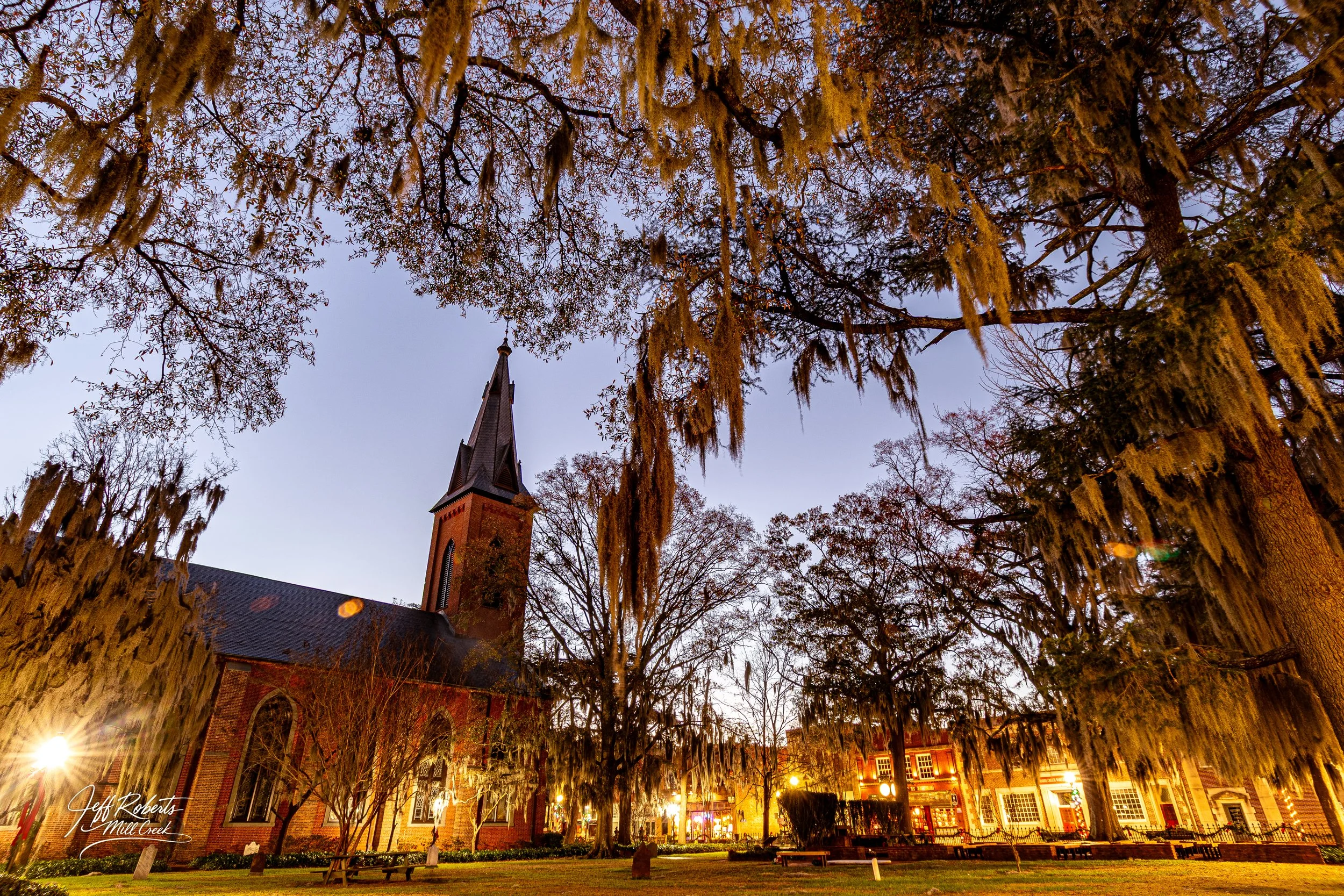 Church with a tall steeple surrounded by large trees with hanging Spanish moss in a town park during evening.