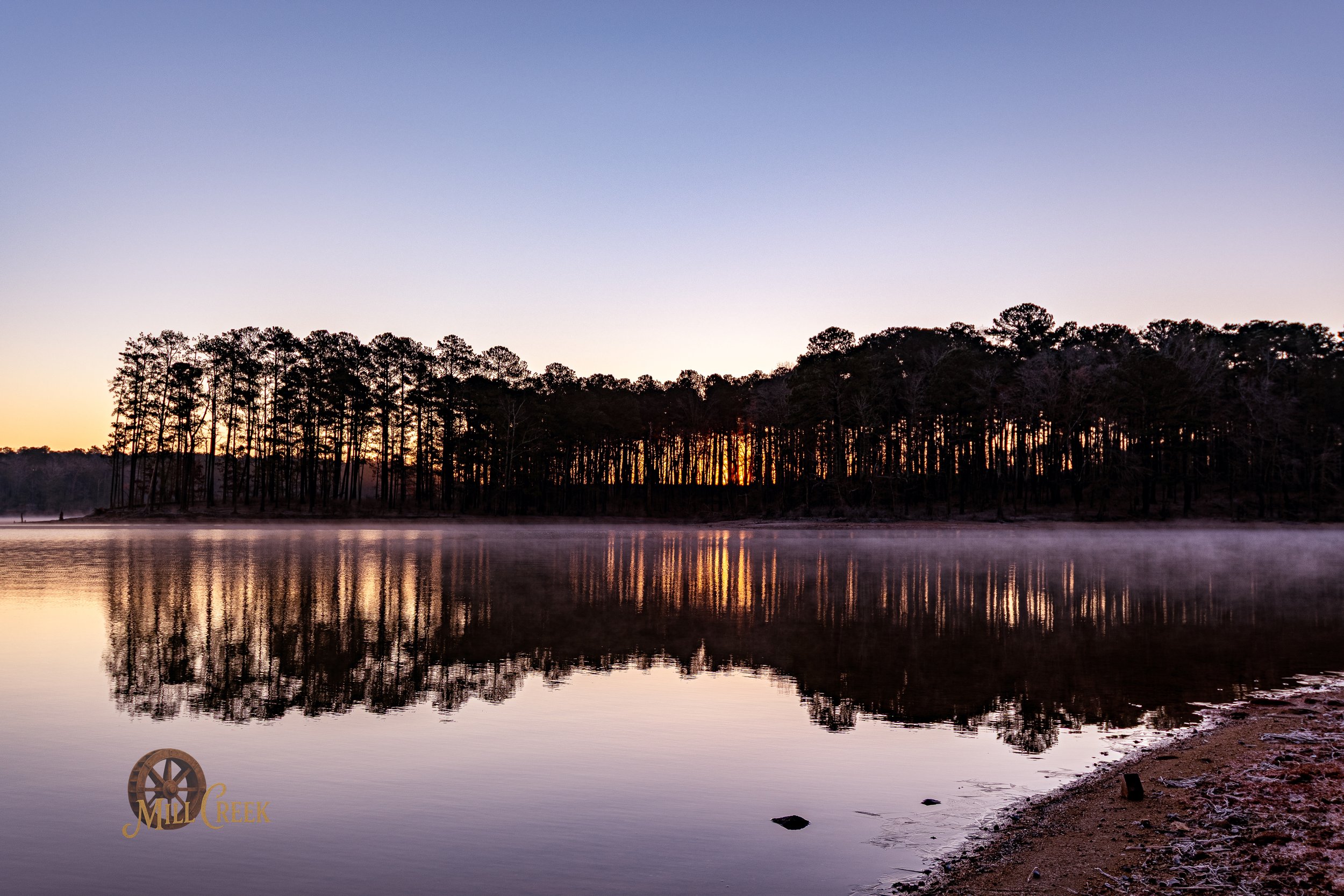 Sunset over a calm lake with a reflection of trees on the water and light fog near the shoreline, with the logo Mill Creek in the bottom left corner.