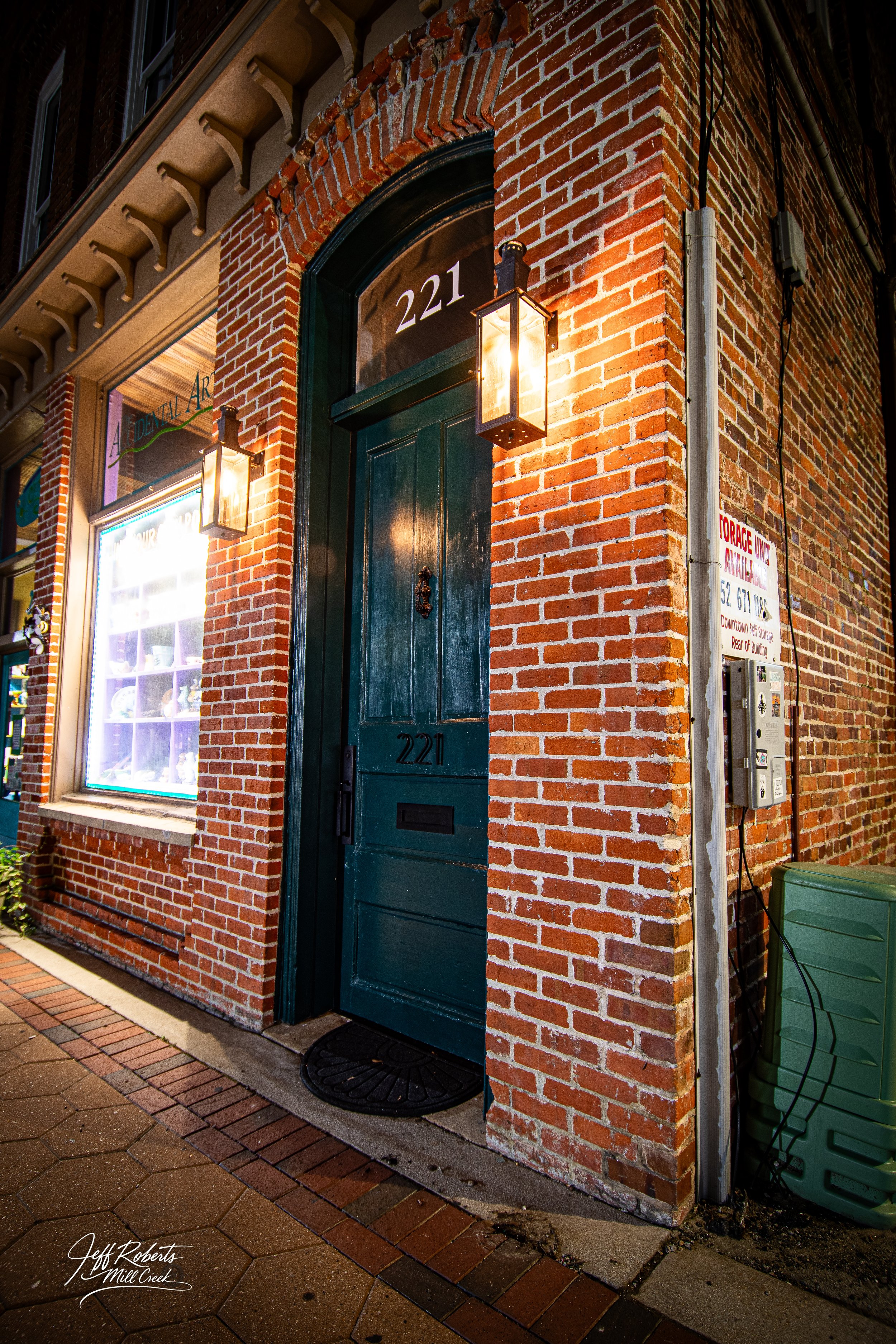 Nighttime view of a brick building with a dark green door marked '221', illuminated by two wall-mounted lanterns. A display window on the left highlights some items, and a sign on the right indicates parking regulations.