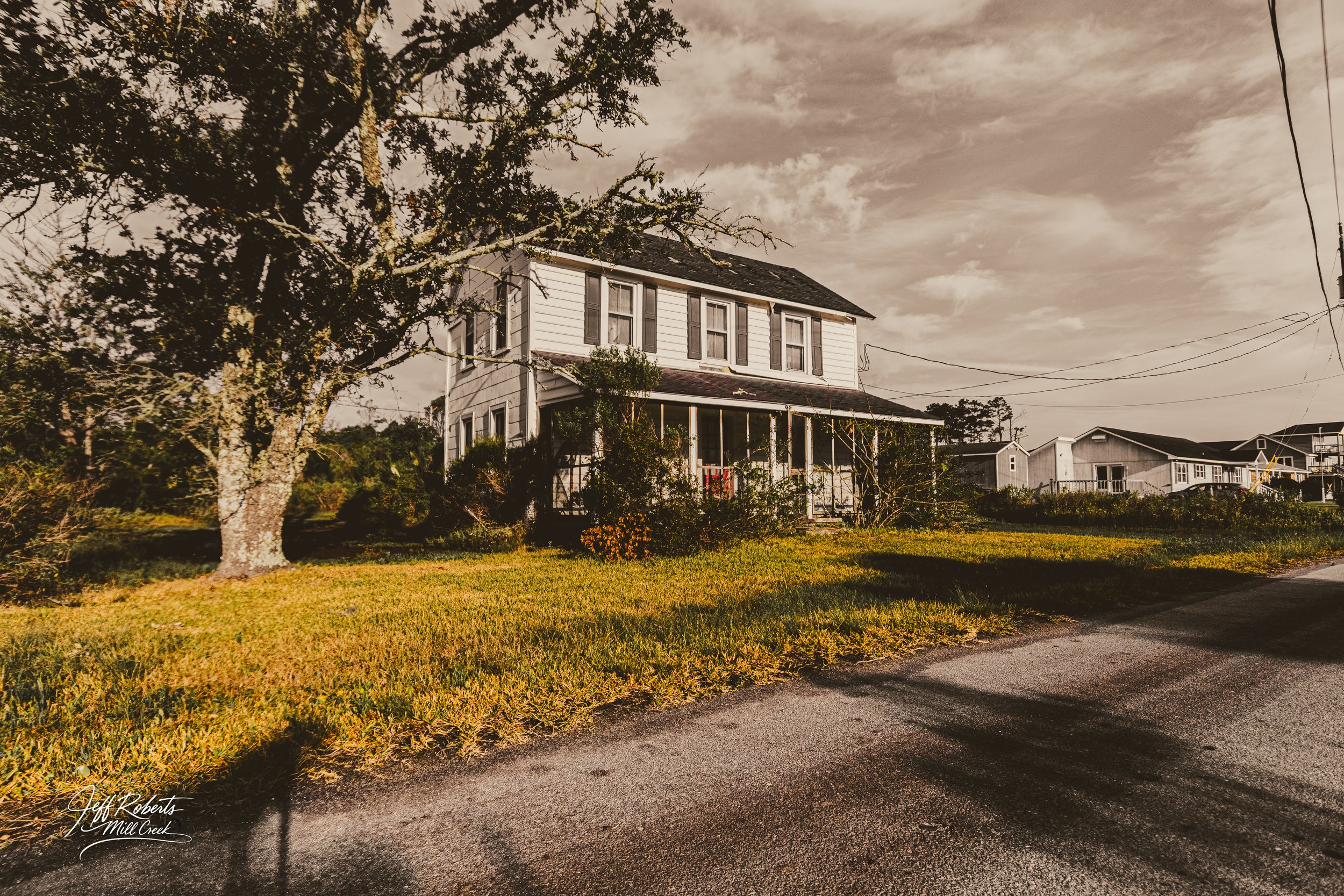 A two-story white house with porch, surrounded by trees and grass, with a cloudy sky overhead.