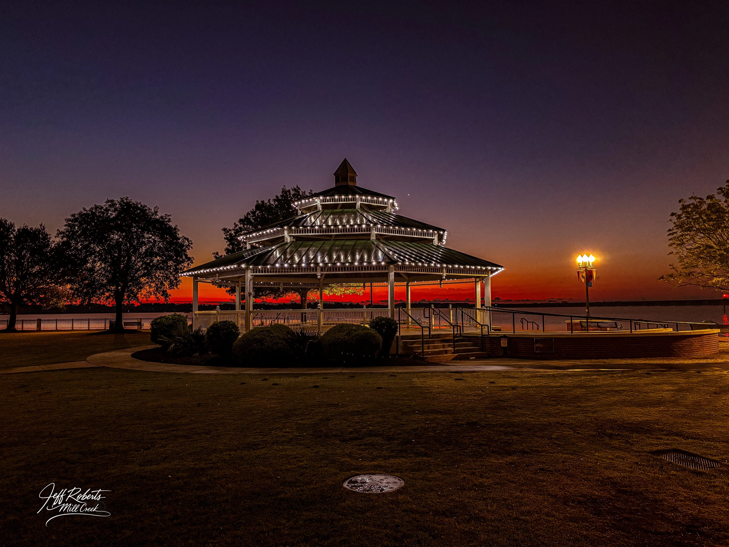 A gazebo with string lights is illuminated at sunset near a body of water, with trees and a lamp post nearby.