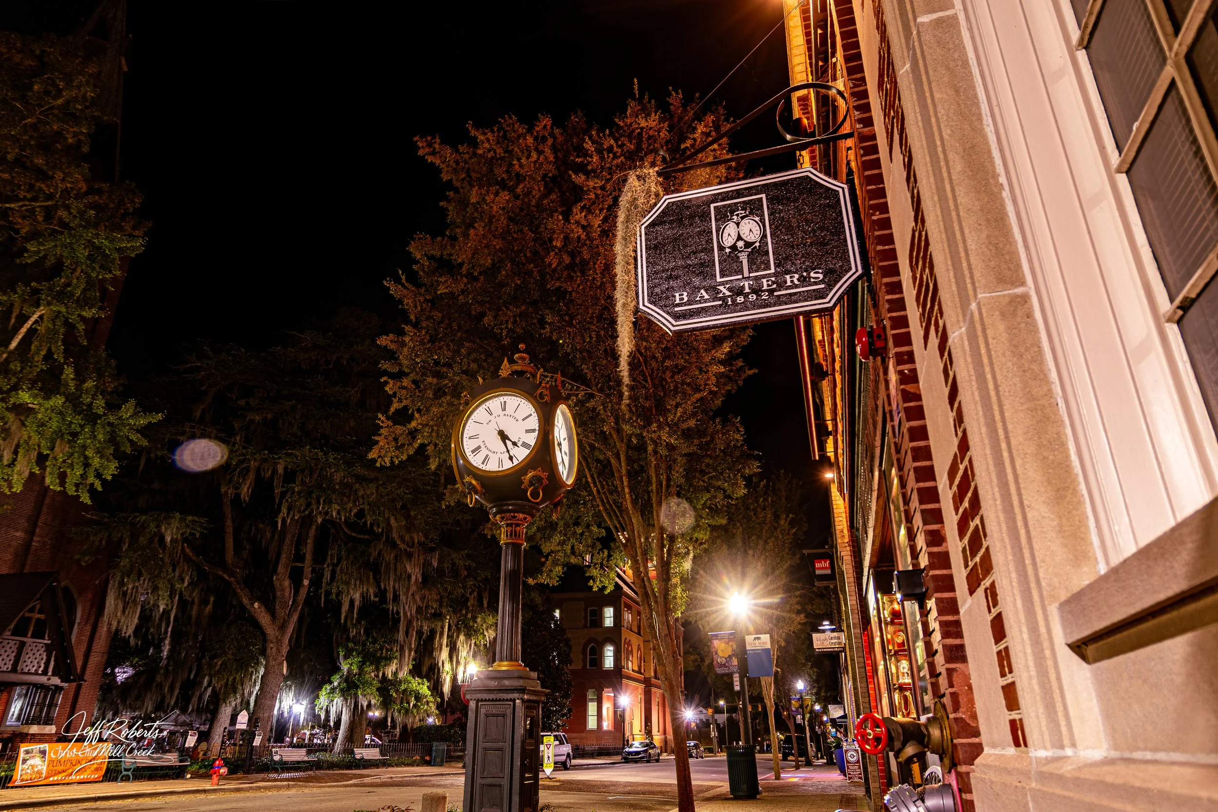 Night scene of a street corner with a vintage clock and hanging sign for Baxter's, established in 1892, illuminated by streetlights. Old brick building with white window trim and fire hydrant in the foreground, trees with hanging moss in the backgrou