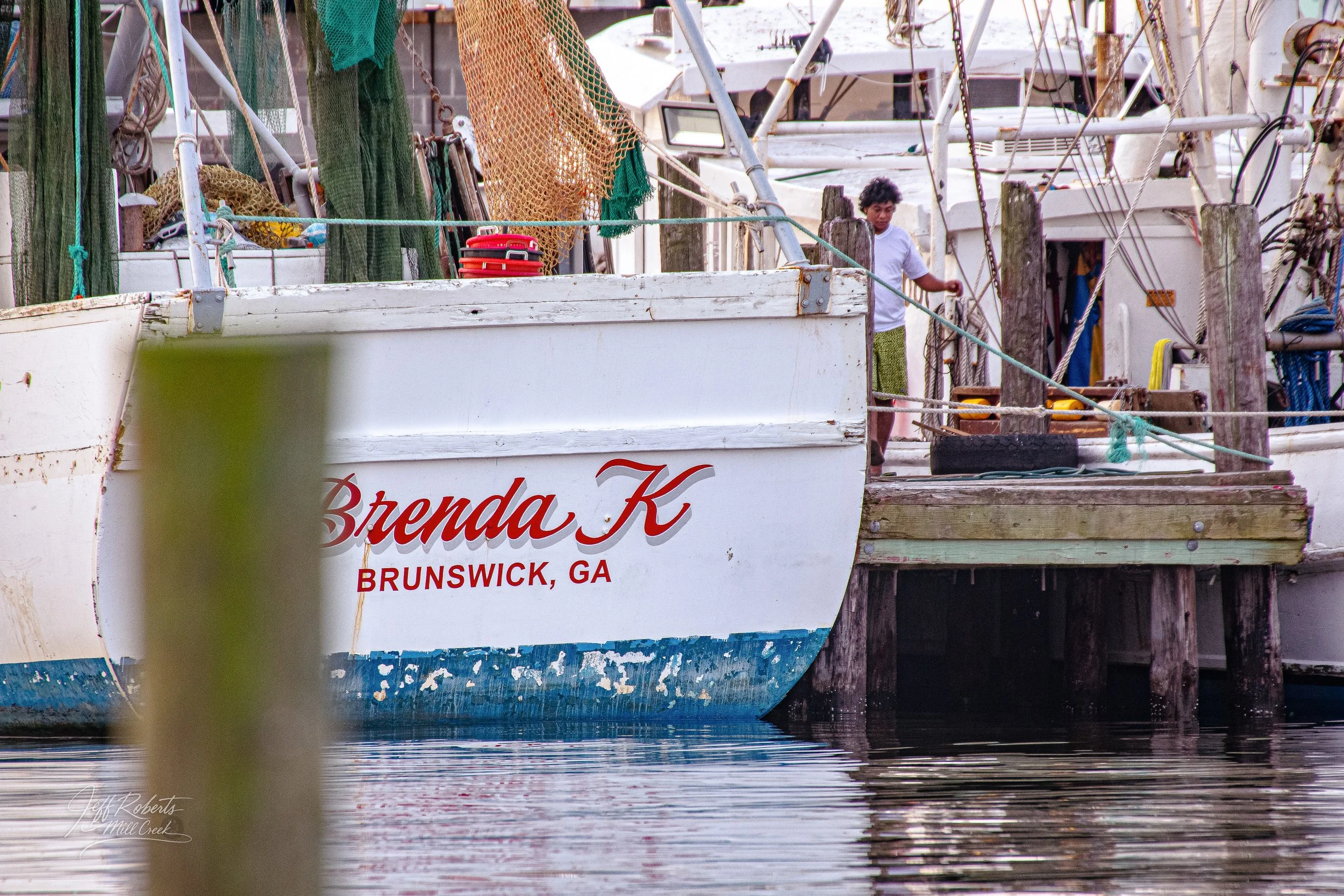 A boat named Brenda K docked at a wooden pier in Brunswick, Georgia. The boat's hull shows signs of wear and peeling paint. A young man with curly hair wearing a white shirt is on the deck near the posts and ropes.
