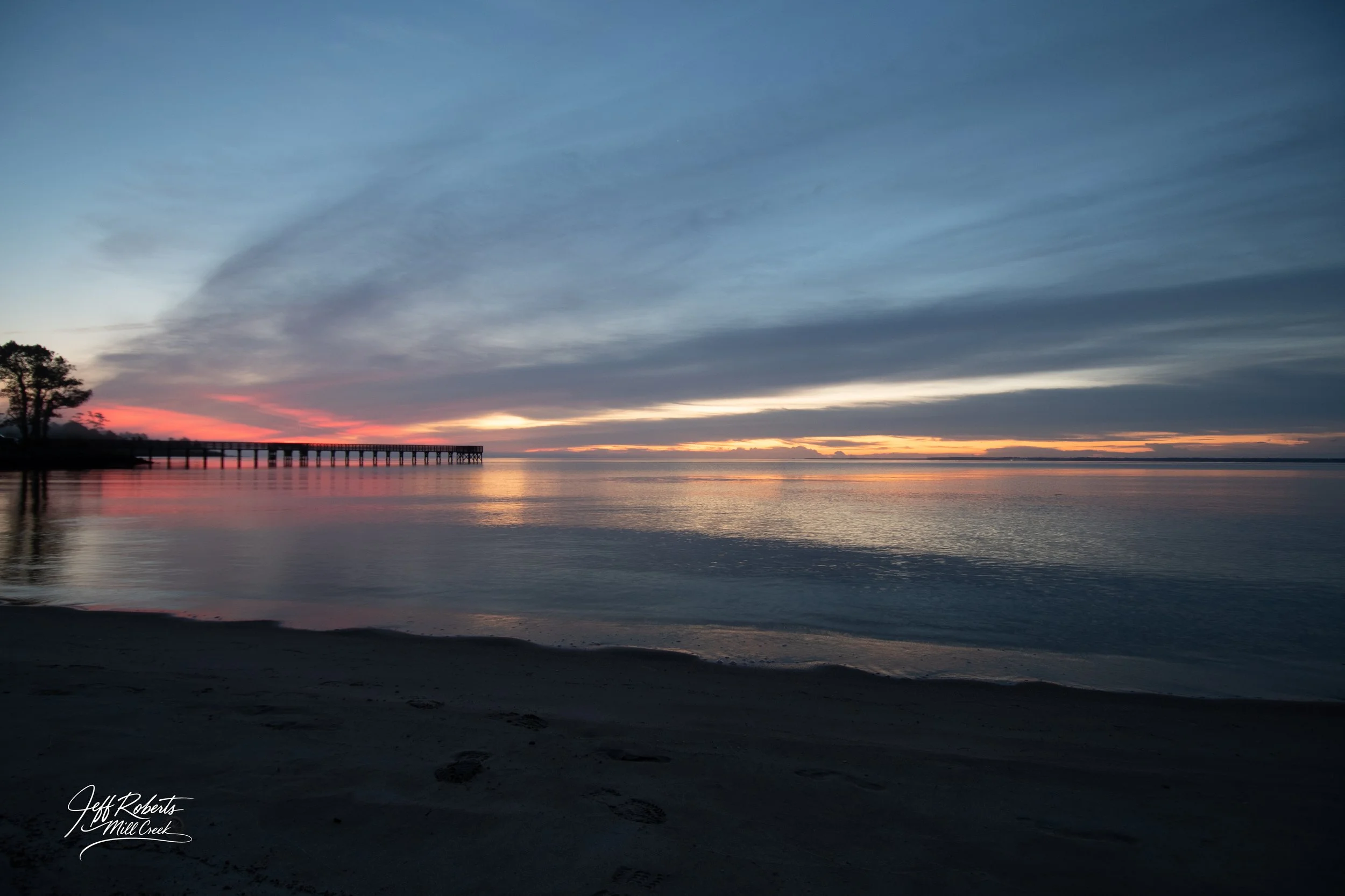 Sunset over a calm body of water with a pier on the left, silhouettes of trees, and footprints in the sandy foreground.