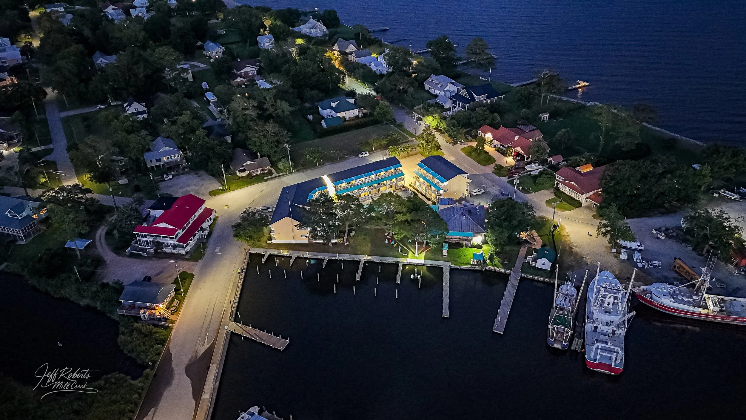 Aerial nighttime view of a marina with boats docked, surrounded by houses along the water's edge, with some houses illuminated, and a small road leading into the area.