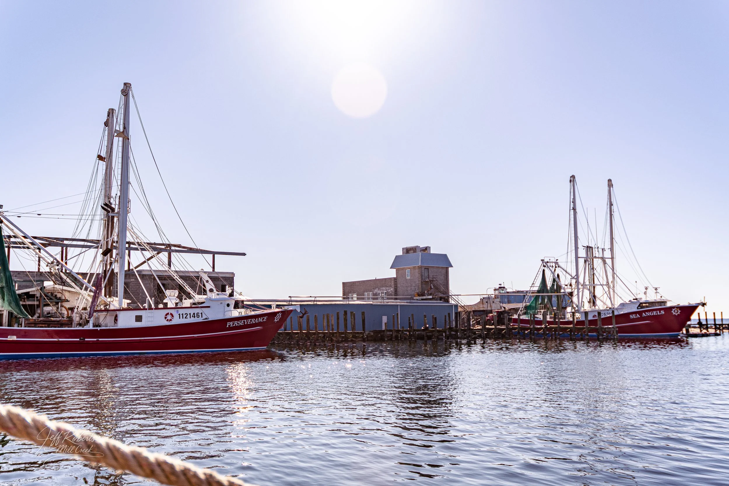 Two red sailboats docked at a harbor with a blue sky and the sun shining overhead.