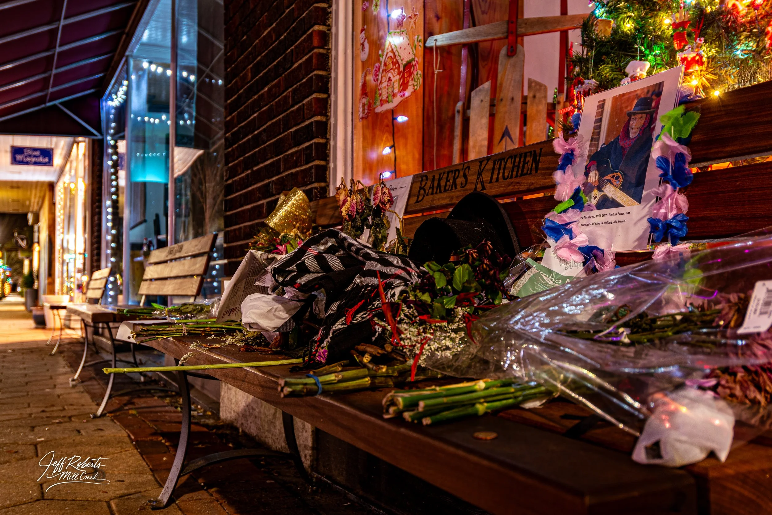 A memorial on a bench with flowers, photos, and signs. Christmas decorations and lights are visible in the background.