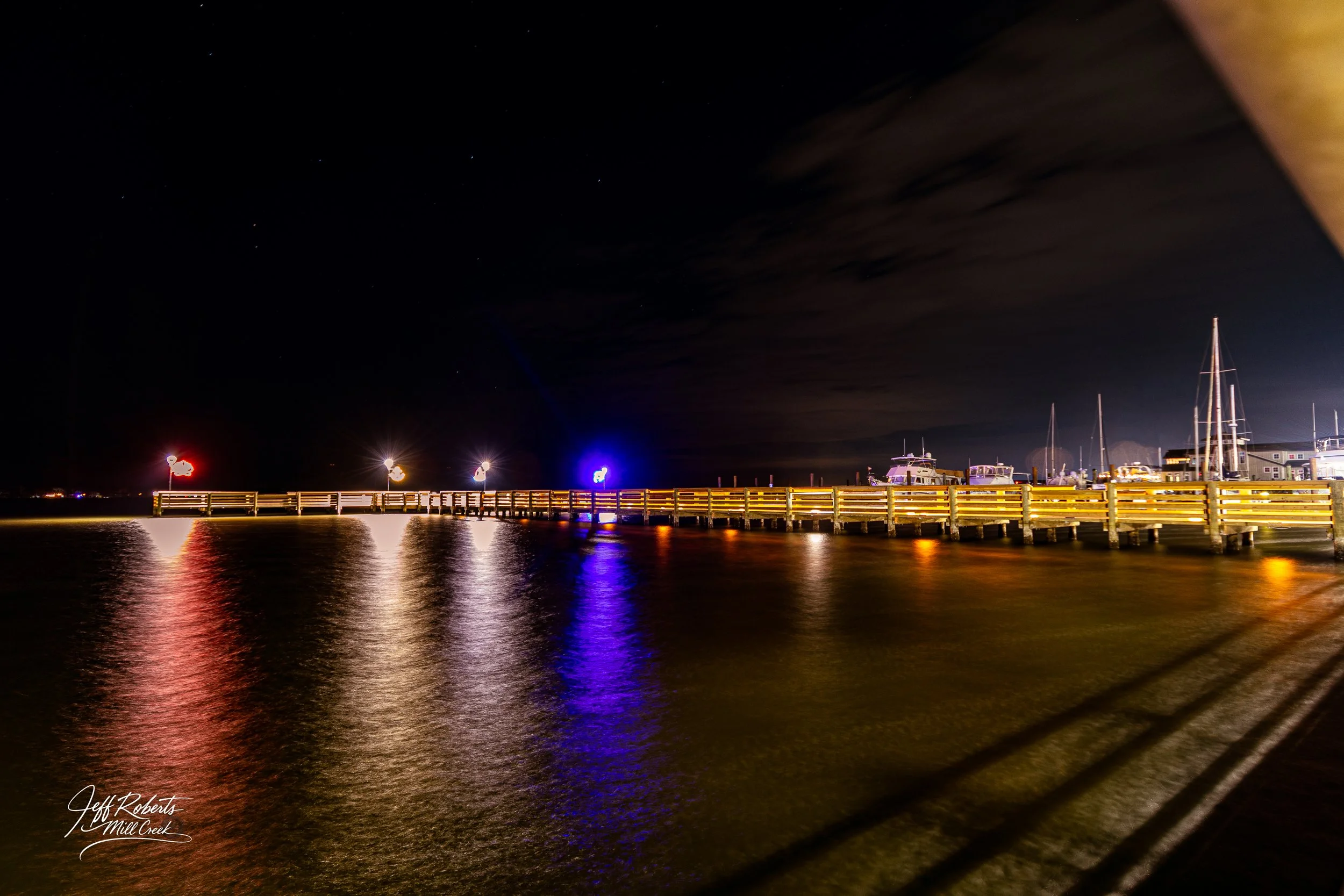 Night view of a wooden pier with colorful lights reflecting on the water, boats docked at the marina, and a cloudy night sky.
