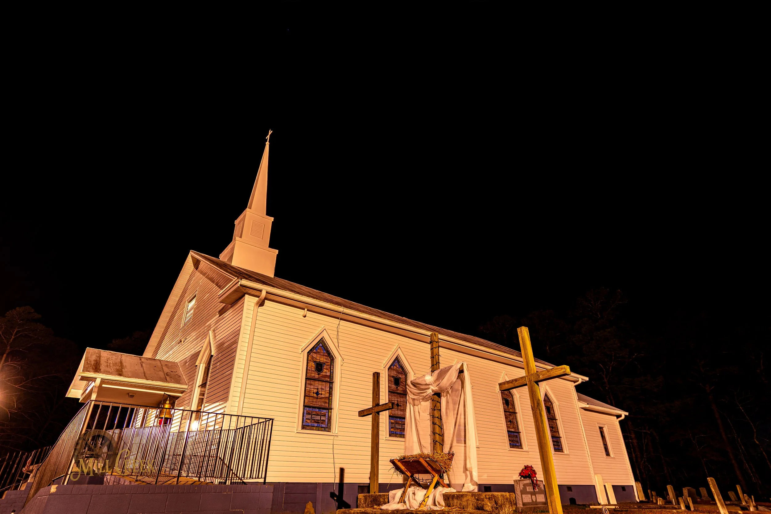 A white church with a tall steeple illuminated at night, featuring stained glass windows and a historic Christian nativity scene with crosses in front.