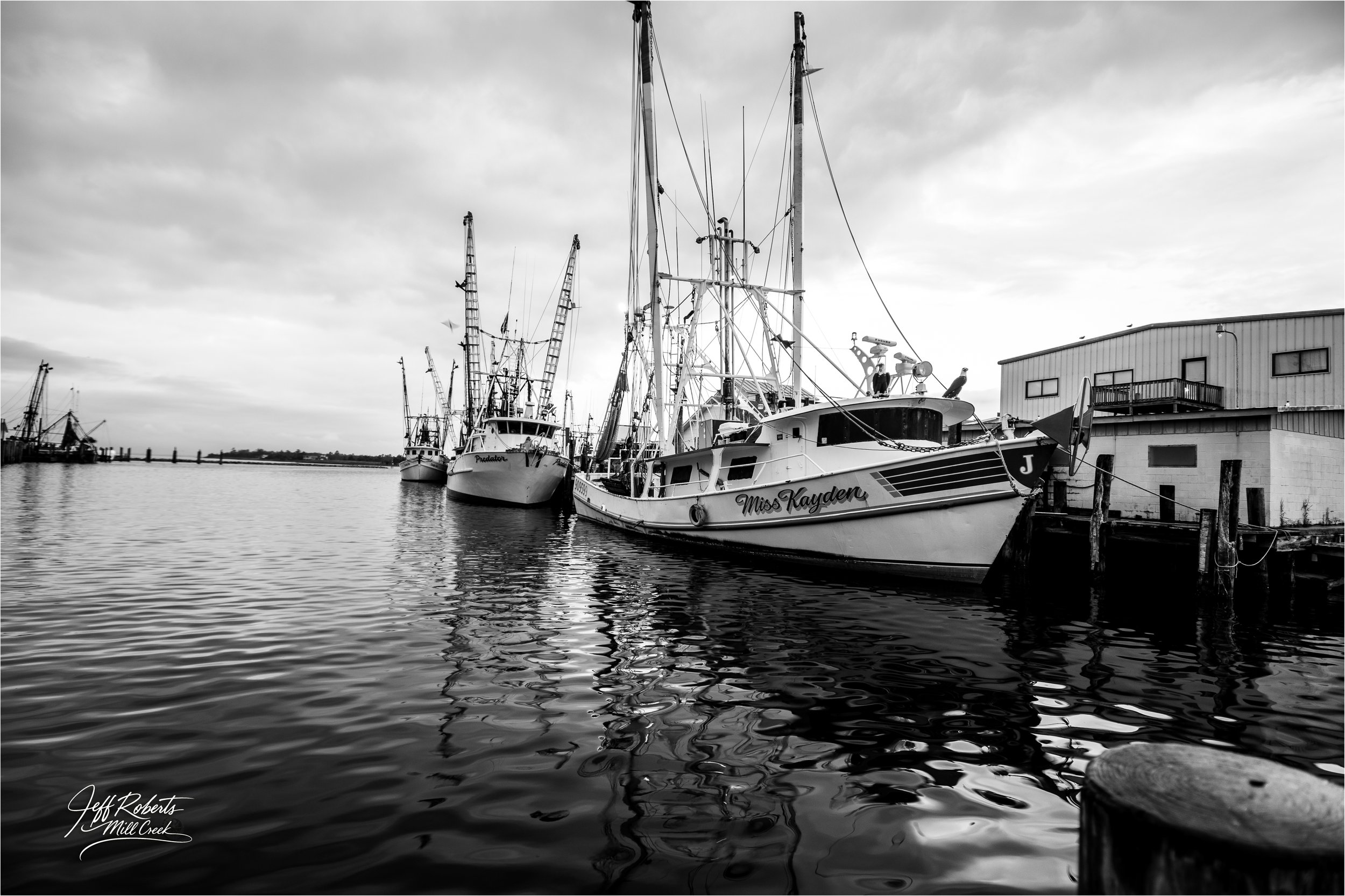 Black and white photo of boats docked at a pier, with a building in the background and calm water reflecting the boats and sky