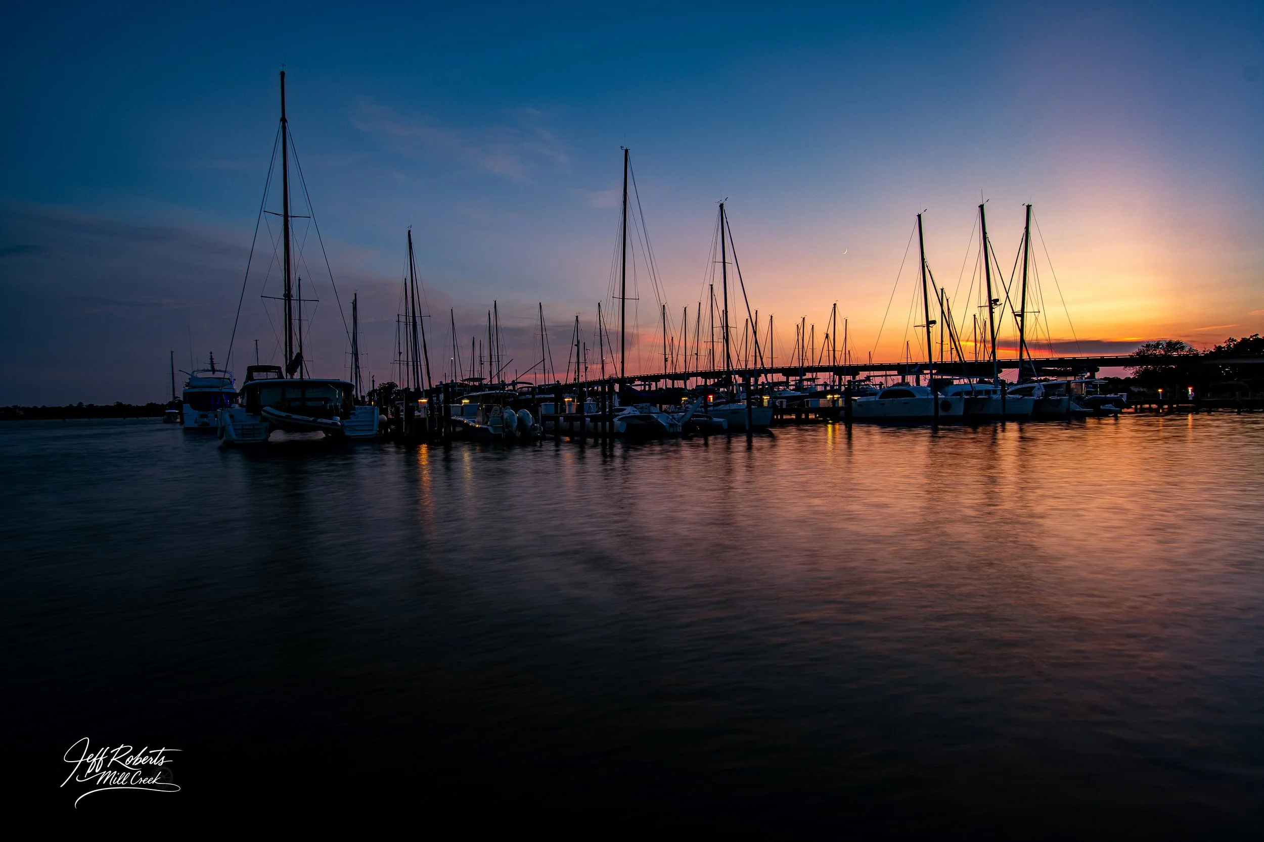 Sailboats and yachts docked at a marina during sunset with colorful sky and water reflecting the sky's colors.
