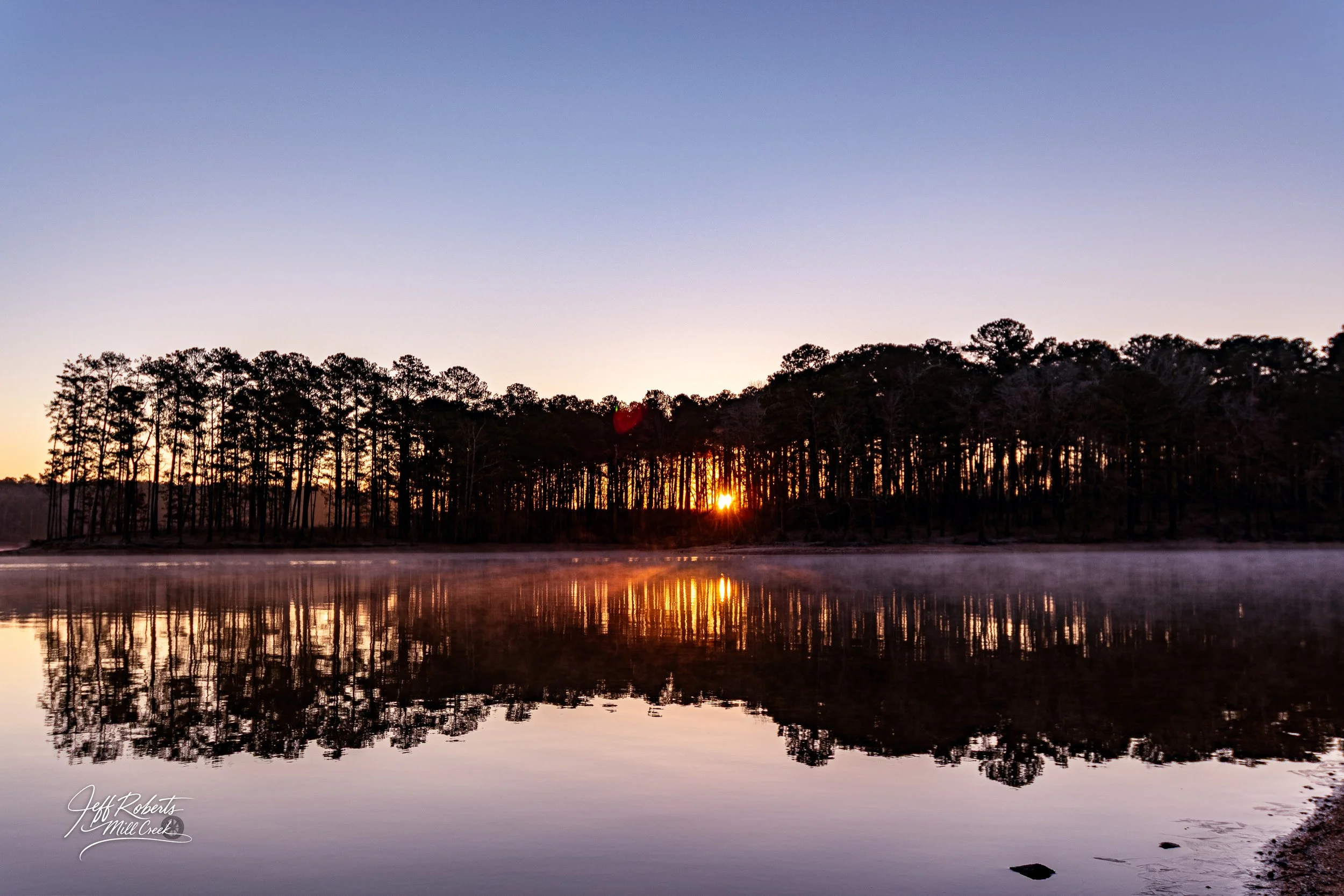 Sunset over a calm lake with a forest of tall pine trees in the background and fog hovering above the water.
