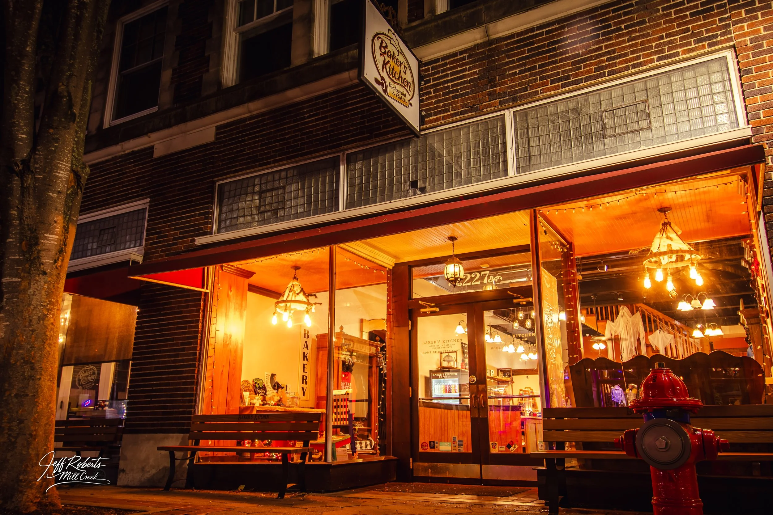 Night view of a bakery and kitchen restaurant with large glass windows and wooden accents, warmly lit with chandeliers and string lights, with benches outside and a red fire hydrant nearby.