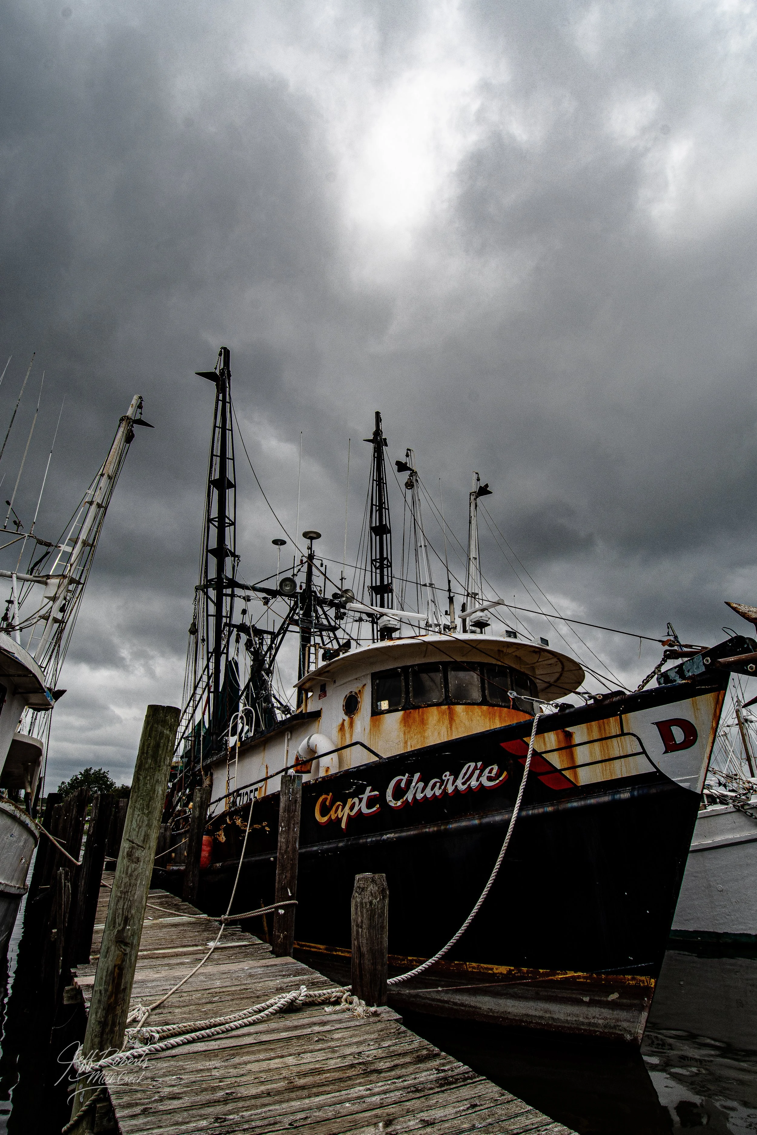 A rusty, black fishing boat named 'Capt Charlie' docked at a wooden pier under a dark, cloudy sky.