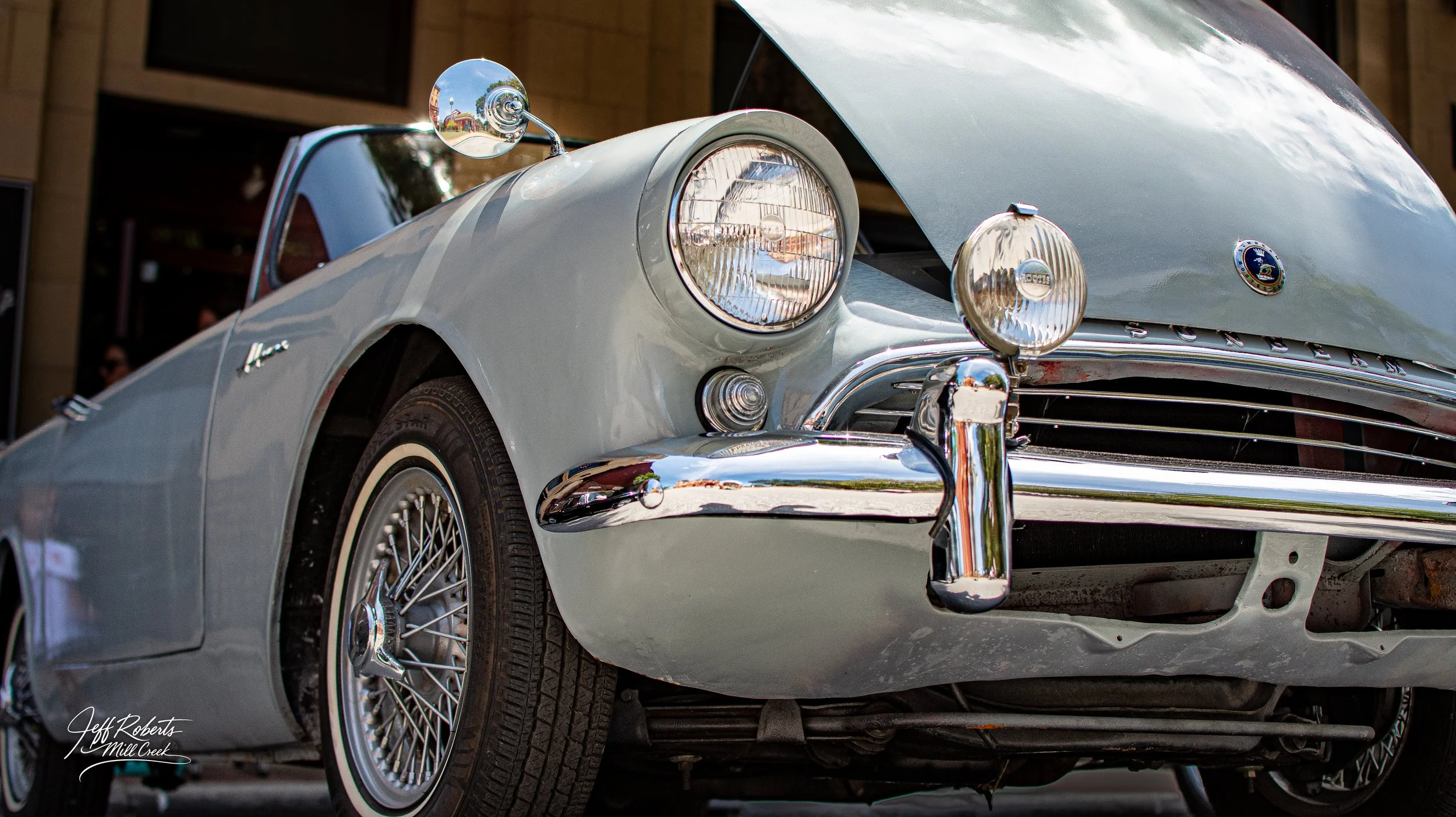 A classic silver Porsche roadster with distinctive round headlights, chrome bumper, and wire spoke wheels on display, with a reflection of the outdoor scene in the mirror.