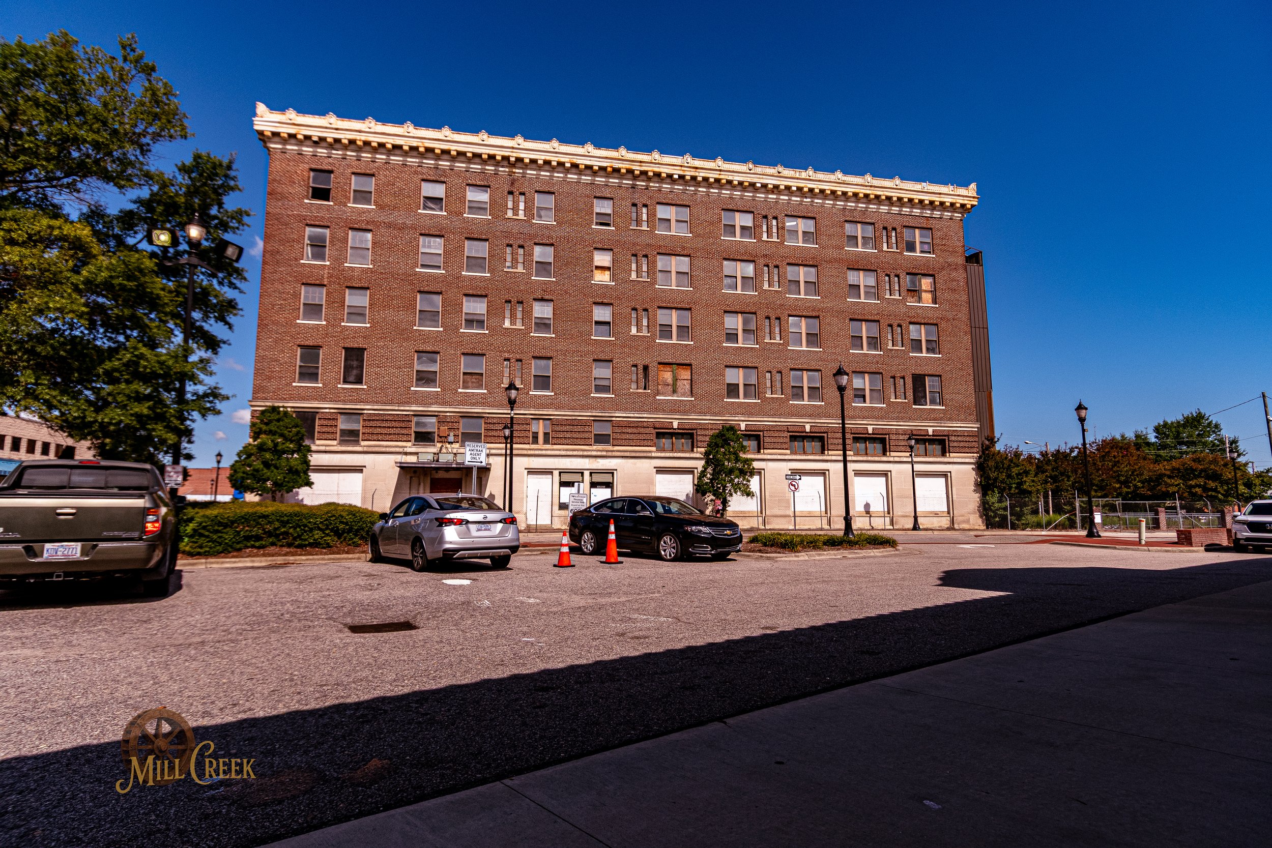 A large brick building with multiple floors, surrounded by parked cars, trees, and street lamps under a clear blue sky.