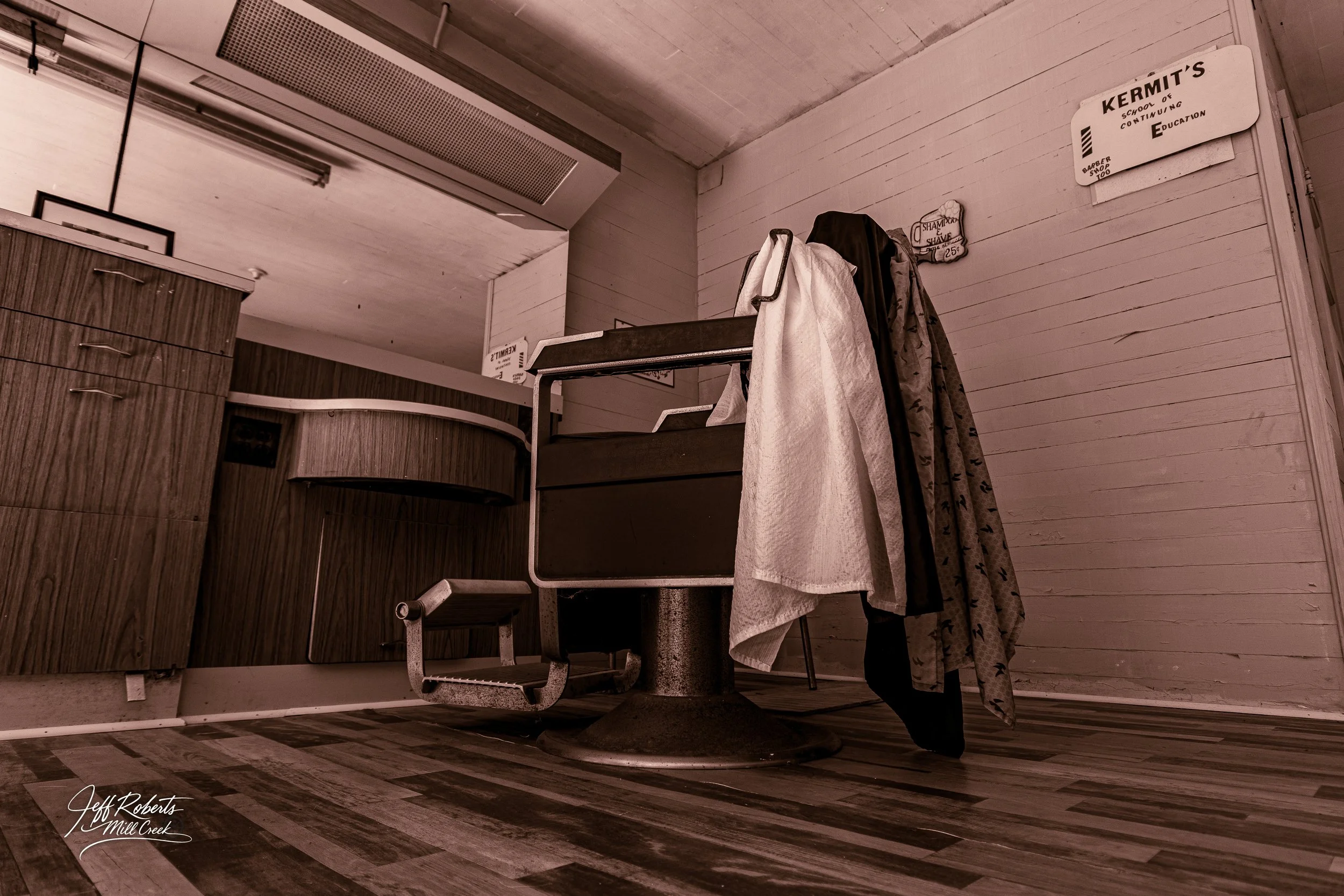 Empty barber shop chair with hanging towels and clothes, wooden cabinets, and a sign on the wall that says "Kermit’s School of Continuing Education."