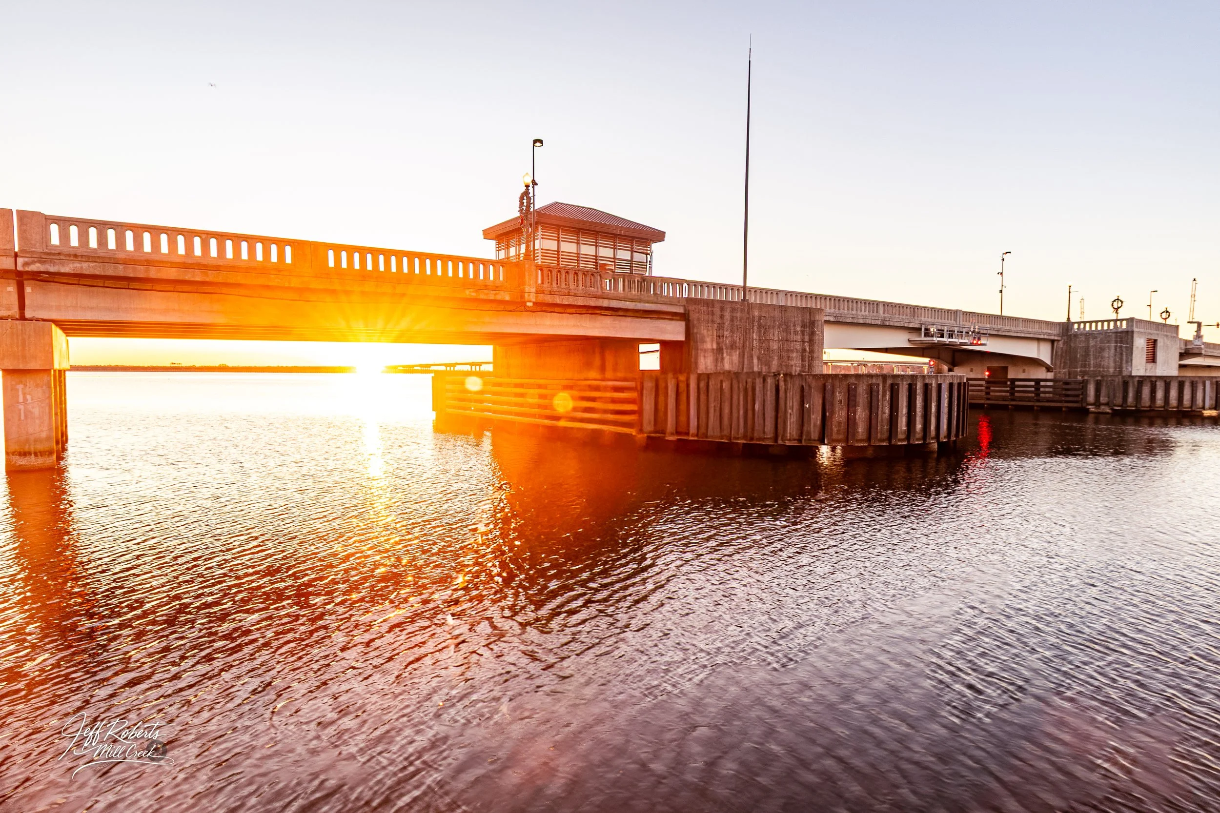 Sunset view of a bridge over water with the sun setting behind it, creating a bright glow and reflections on the water surface.