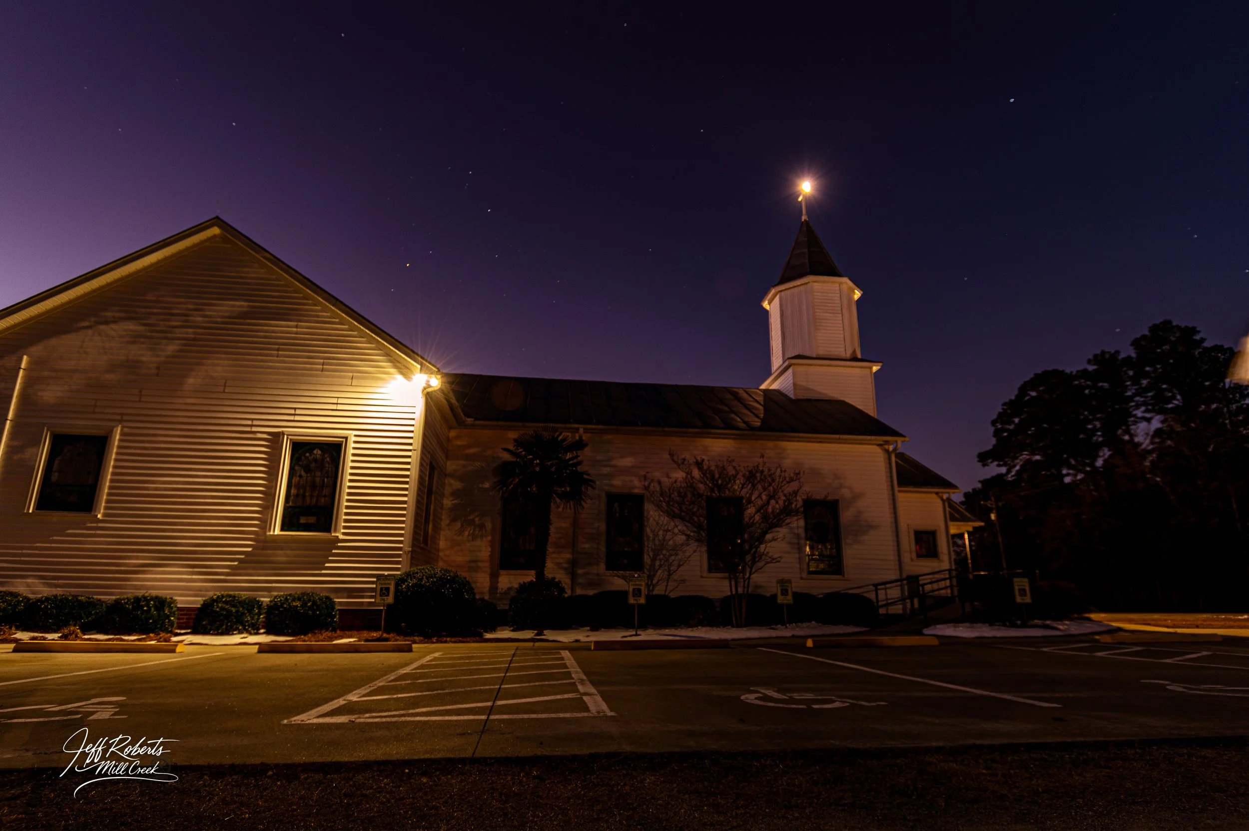 Nighttime view of a church with a steeple, illuminated by exterior lights, surrounded by a parking lot and trees, under a starry sky.