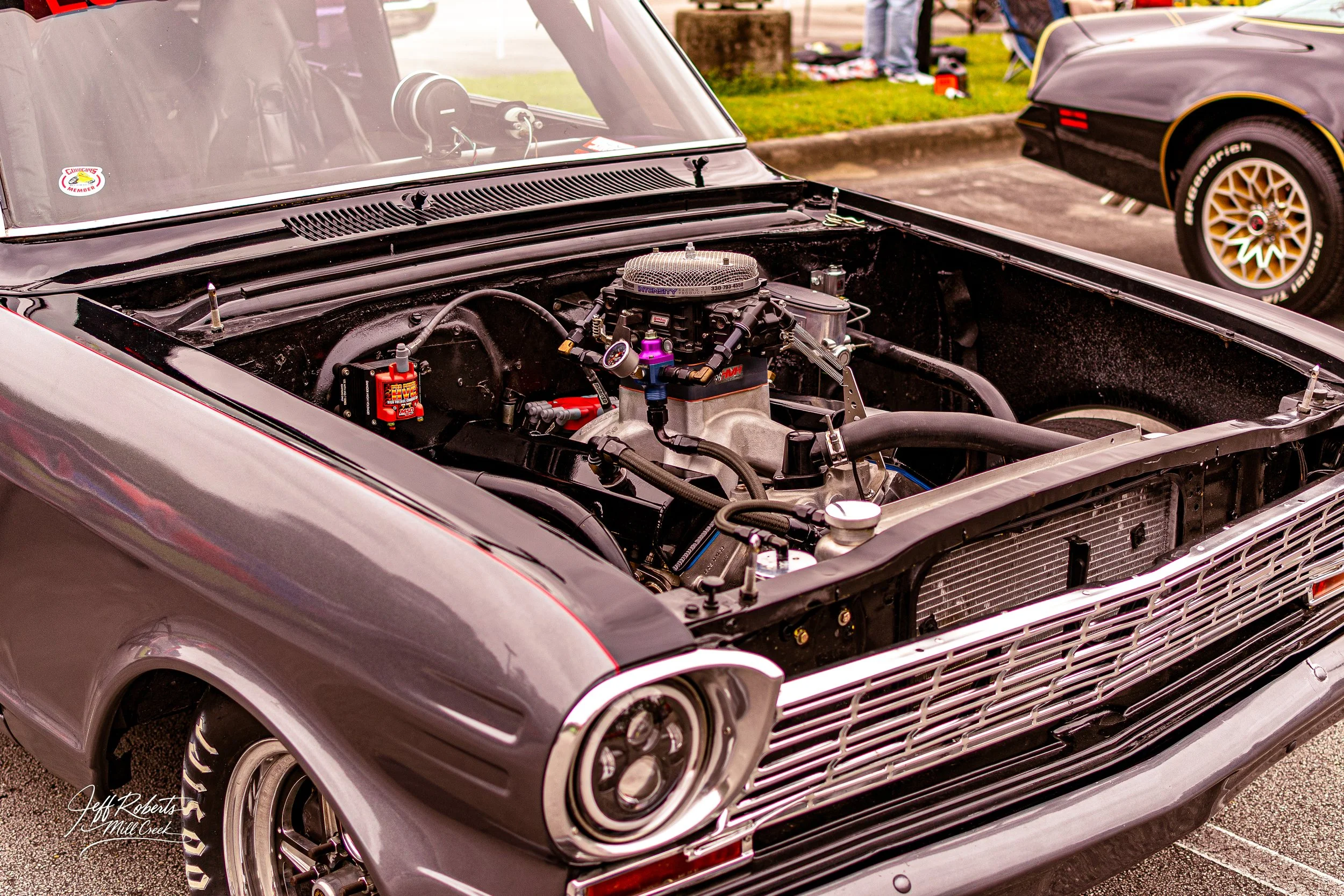 Front view of a vintage black and gray muscle car with its hood removed, exposing the engine, at a car show. Other vehicles and people are visible in the background.