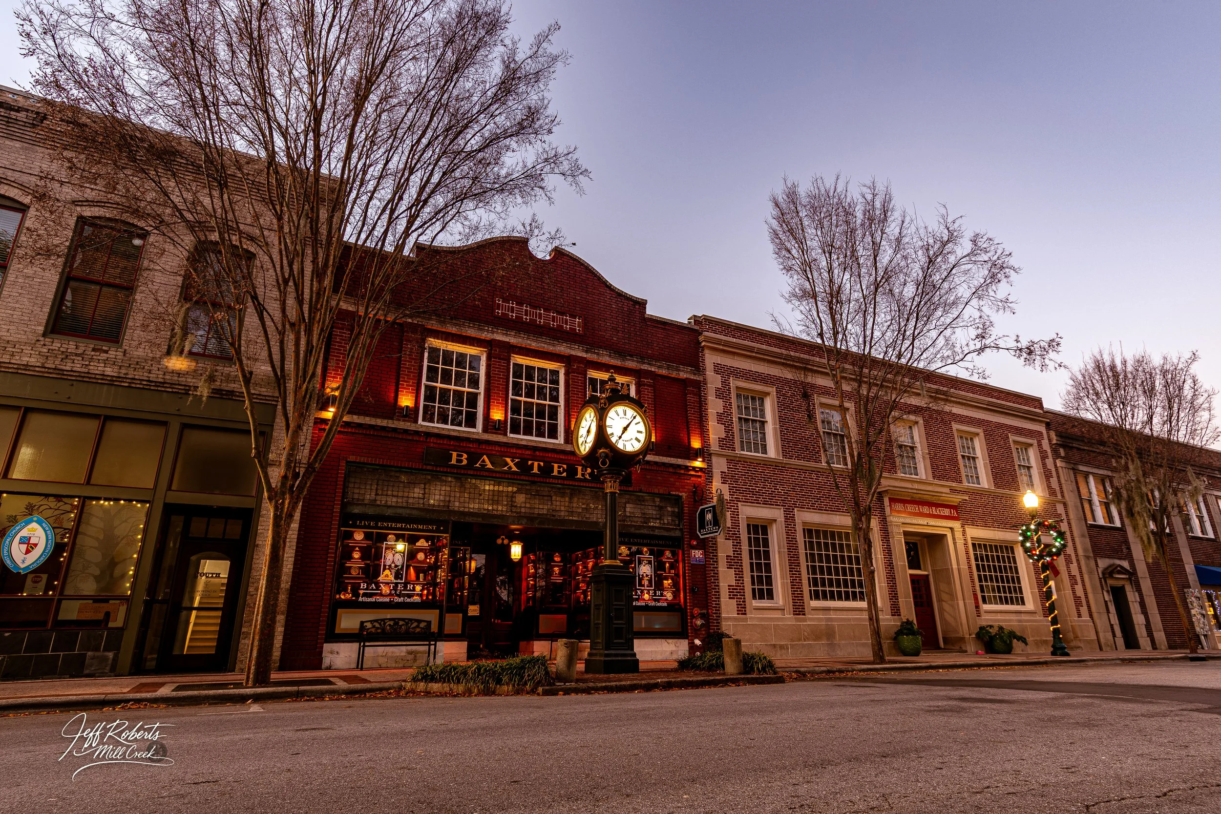 Street view of historic brick buildings at sunset, featuring a clock post in front of a restaurant called Baxter, with festive Christmas decorations on street lamps and leafless trees lining the sidewalk.