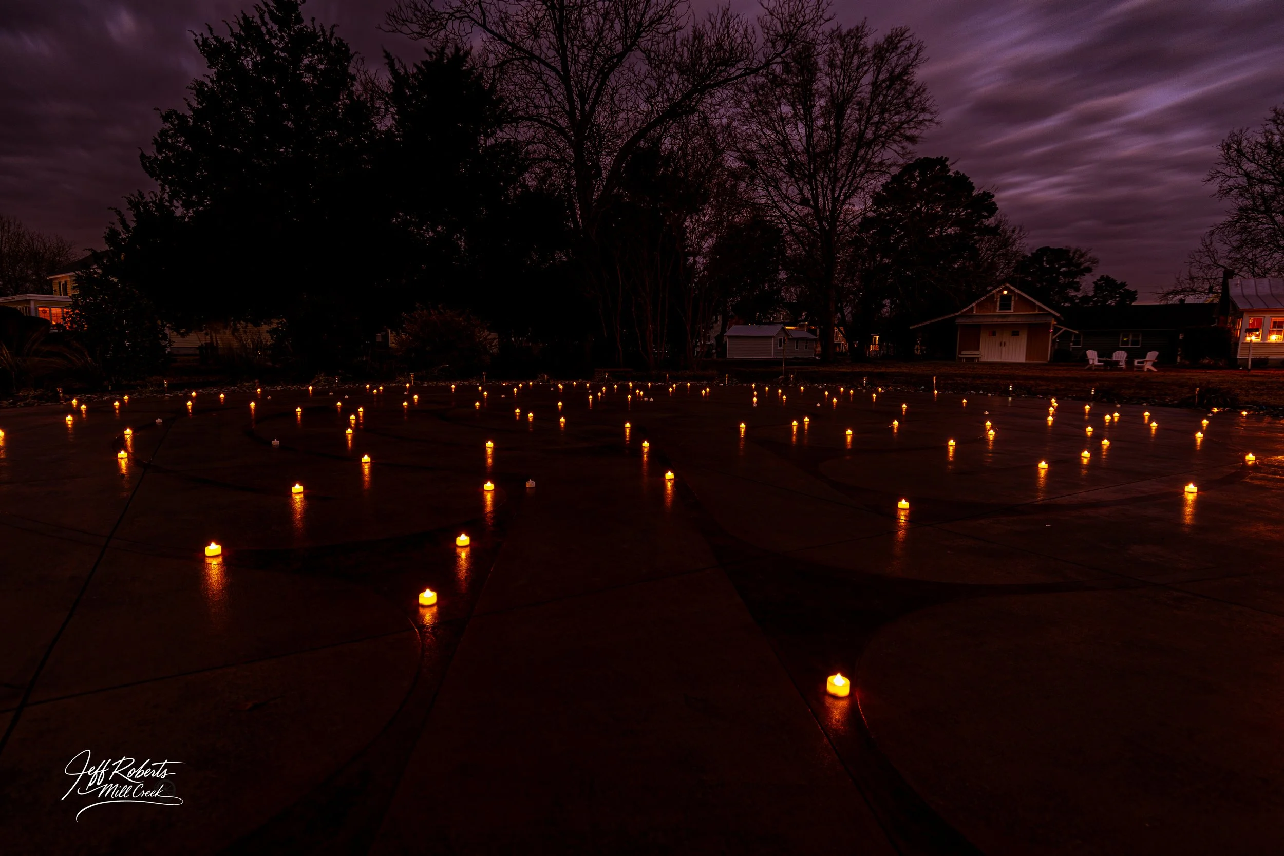 Nighttime scene with numerous candles placed on a wet concrete surface, illuminating the dark surroundings. Silhouettes of trees and houses with lit windows are visible in the background under a purple, cloudy sky.