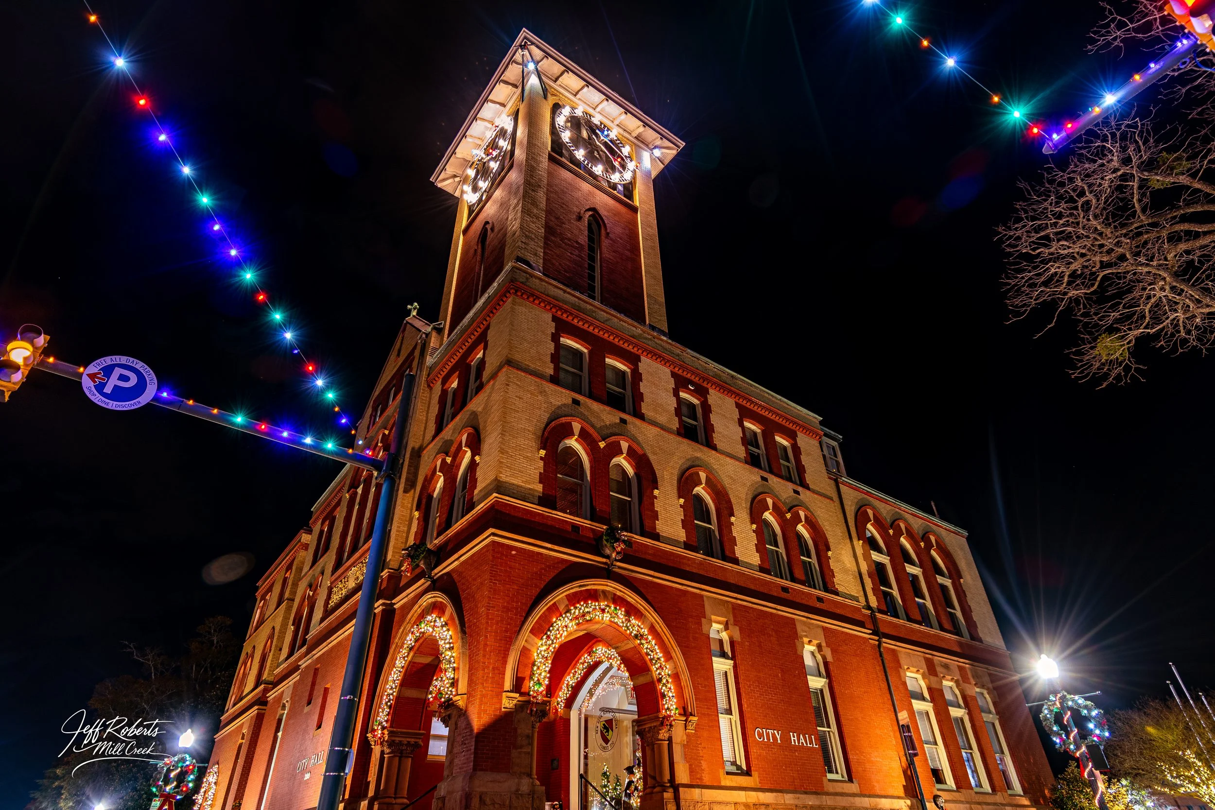 A historic city hall building decorated with Christmas lights and ornaments at night, with colorful string lights hanging above the street.