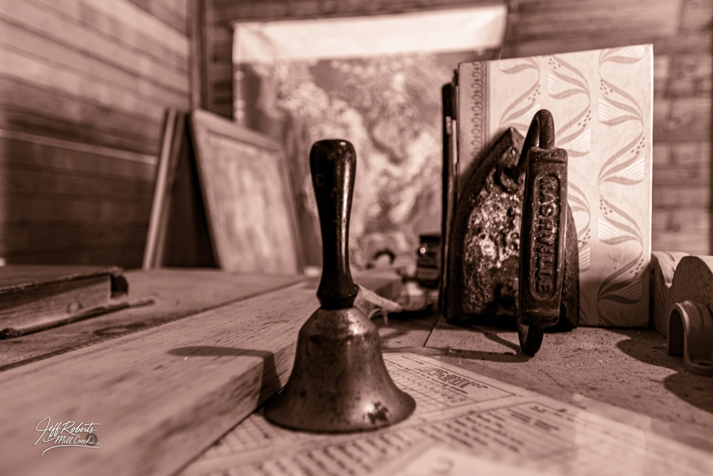 Vintage objects on a wooden surface, including an old bell, a rusty iron, framed photographs, a patterned box, and a sheet of paper with printed text.