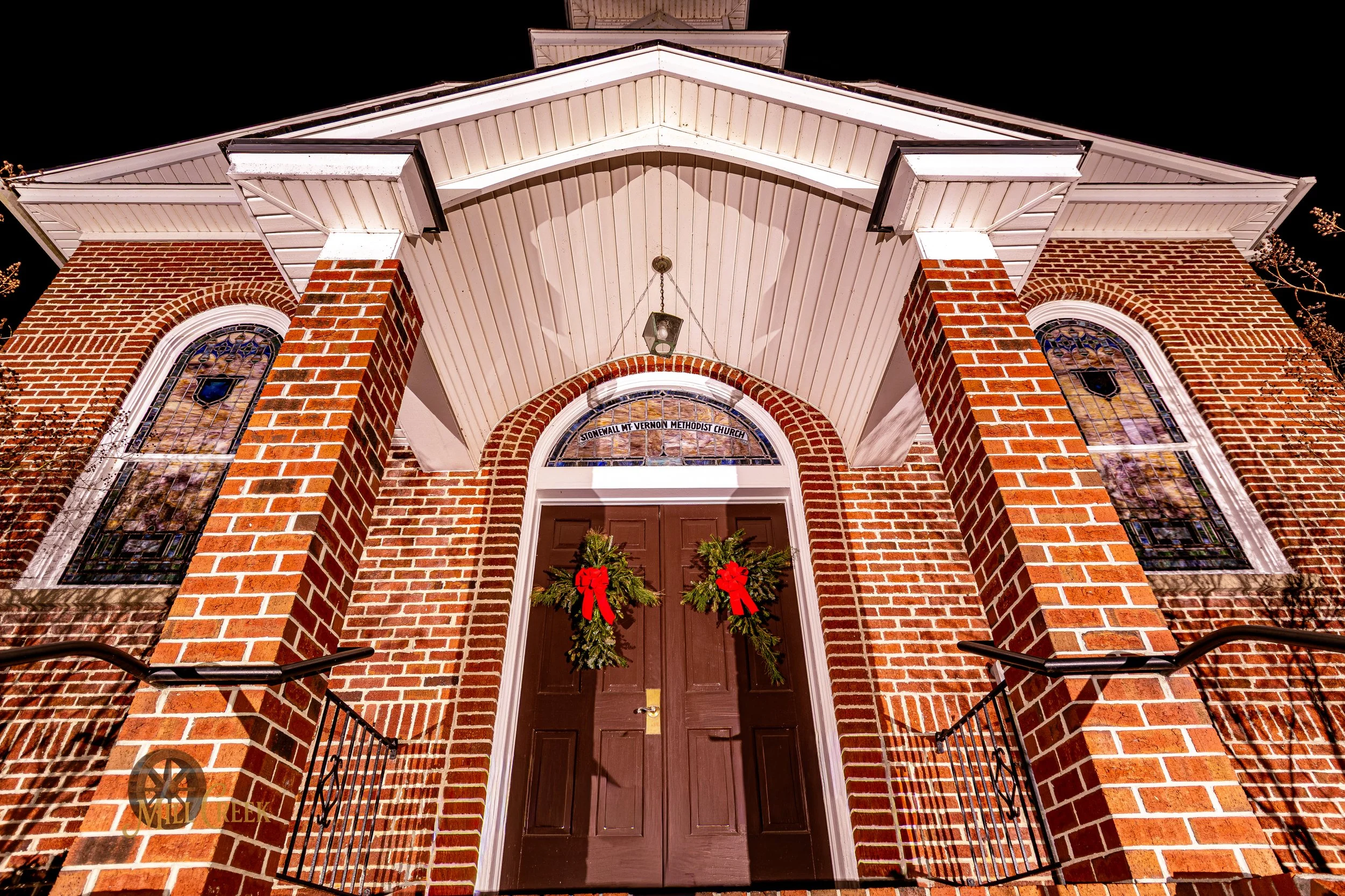 Nighttime view of the brick exterior of a church, with decorated doors and stained glass windows, adorned with Christmas wreaths.