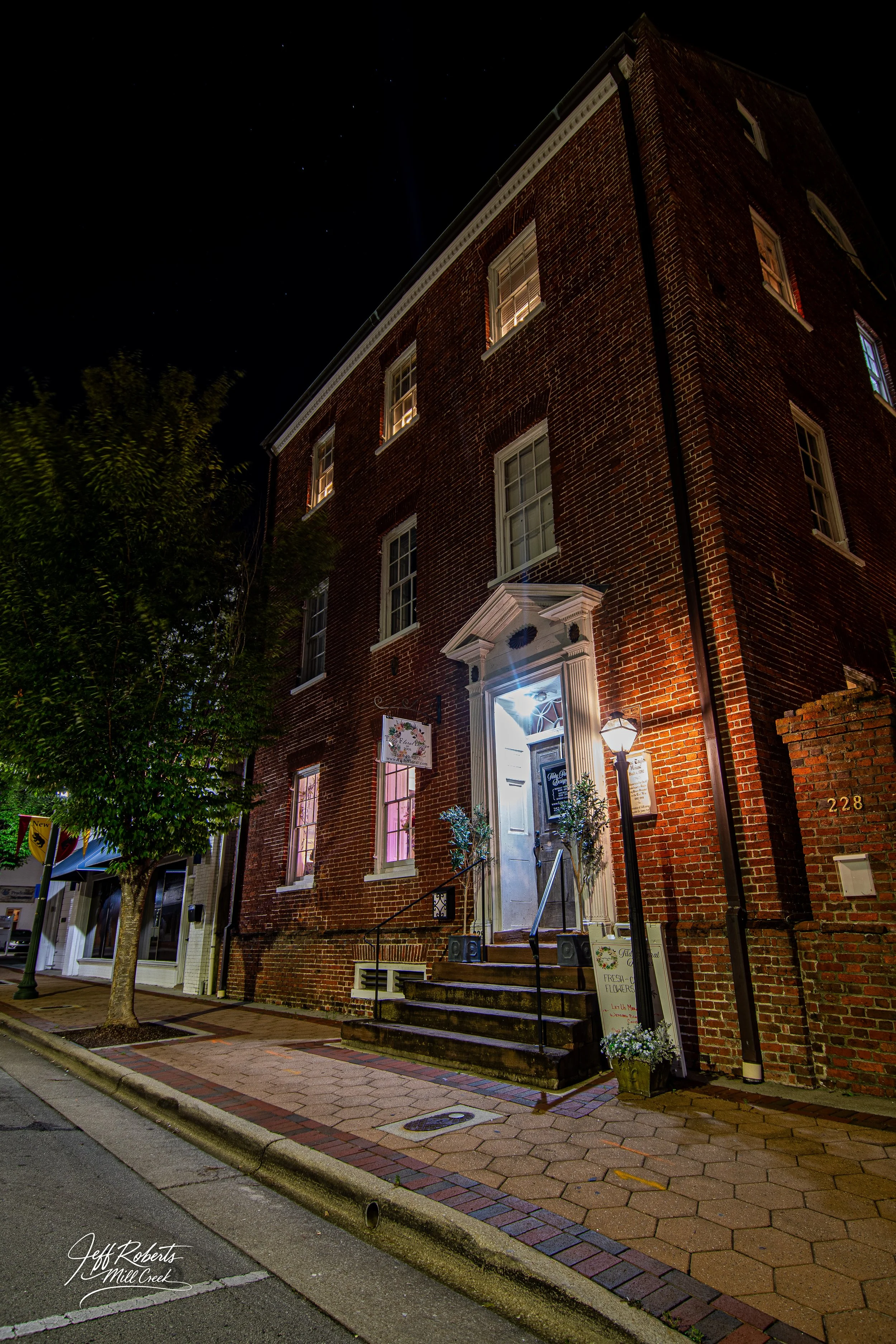 A brick building at night with illuminated door and street lamp, showing steps leading to the entrance, surrounded by sidewalk and trees.