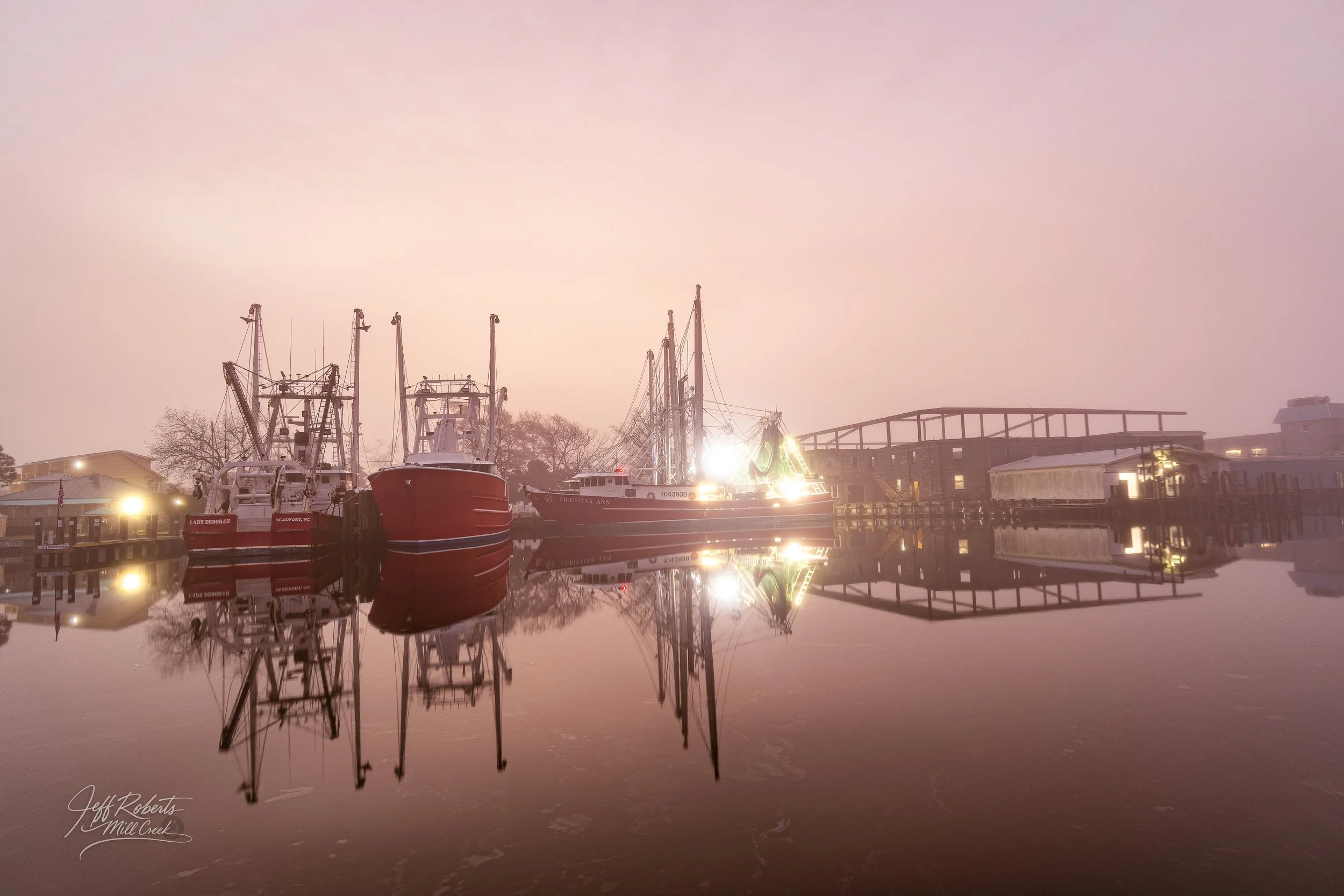 Boats docked at a pier with a foggy sky and calm water reflecting the boats and buildings.