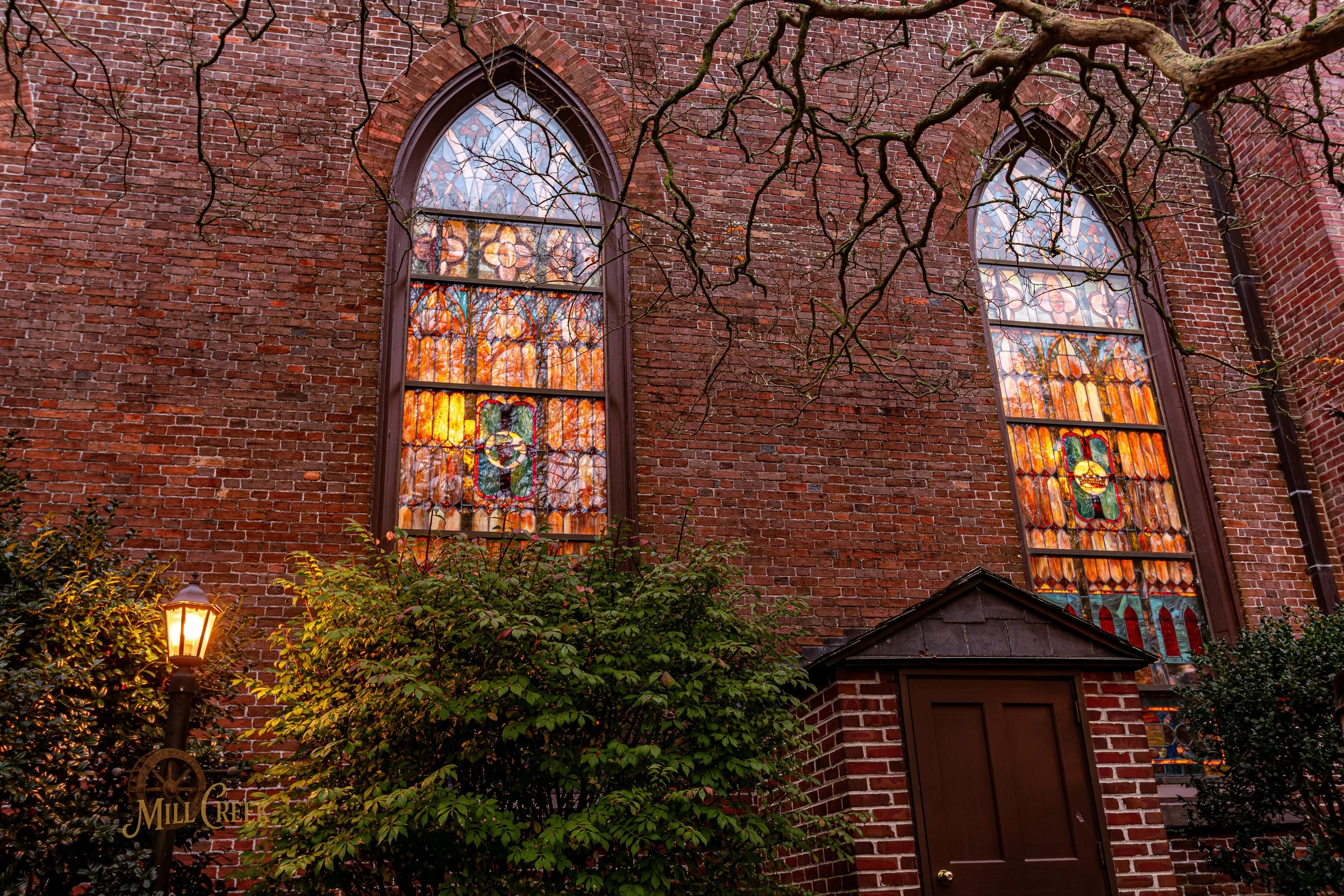 Two tall stained glass windows on a red brick church with leafless tree branches in the foreground, a small brick structure with a door, a lit streetlamp, and bushes.