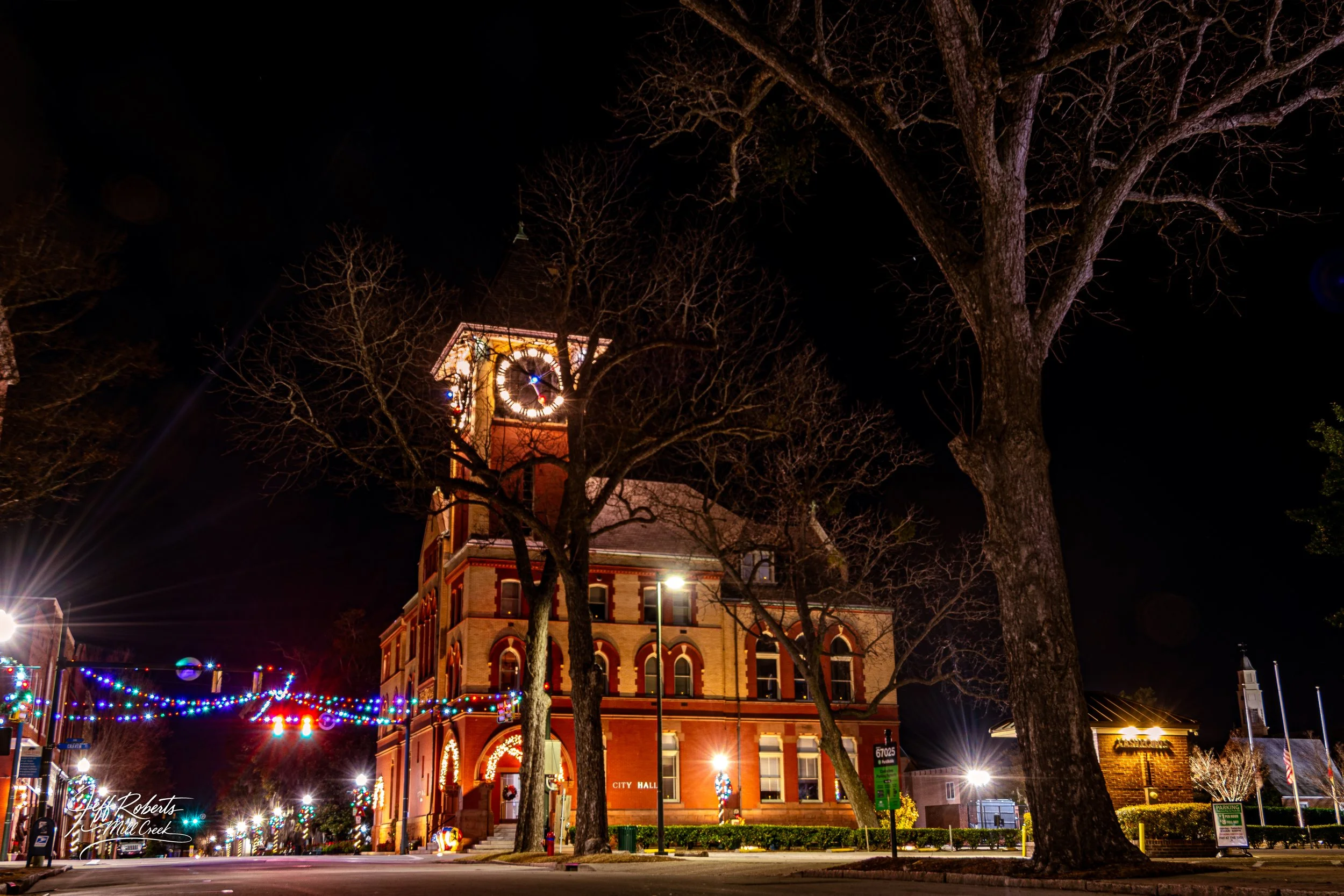 Night view of a historic brick city hall building with a clock tower, decorated with Christmas lights, with leafless trees and streetlights around.