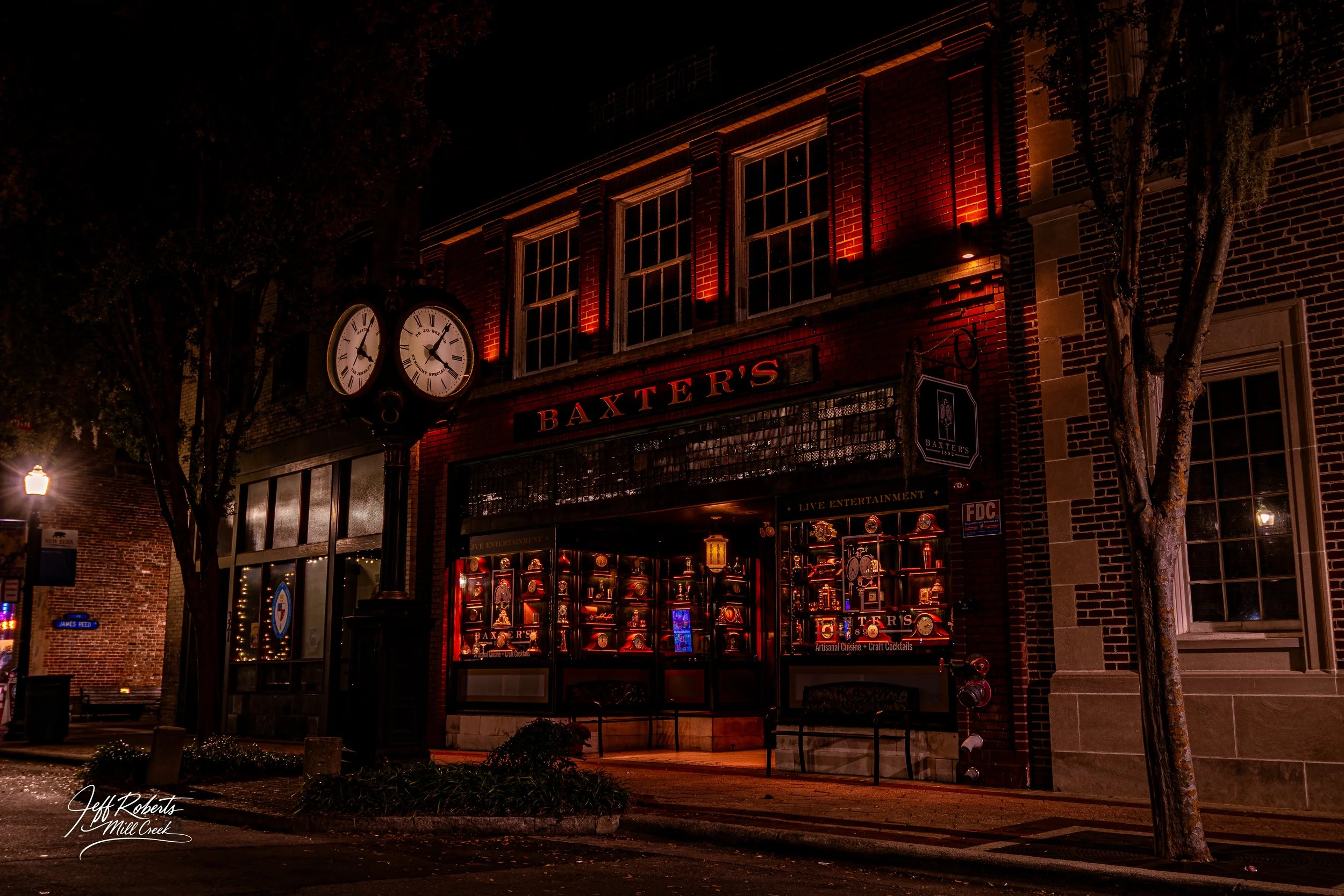 Nighttime exterior view of Baxters, a restaurant or bar, with illuminated red lighting and display windows showcasing bottles and memorabilia, a clock showing approximately 8:10, and trees along the sidewalk.
