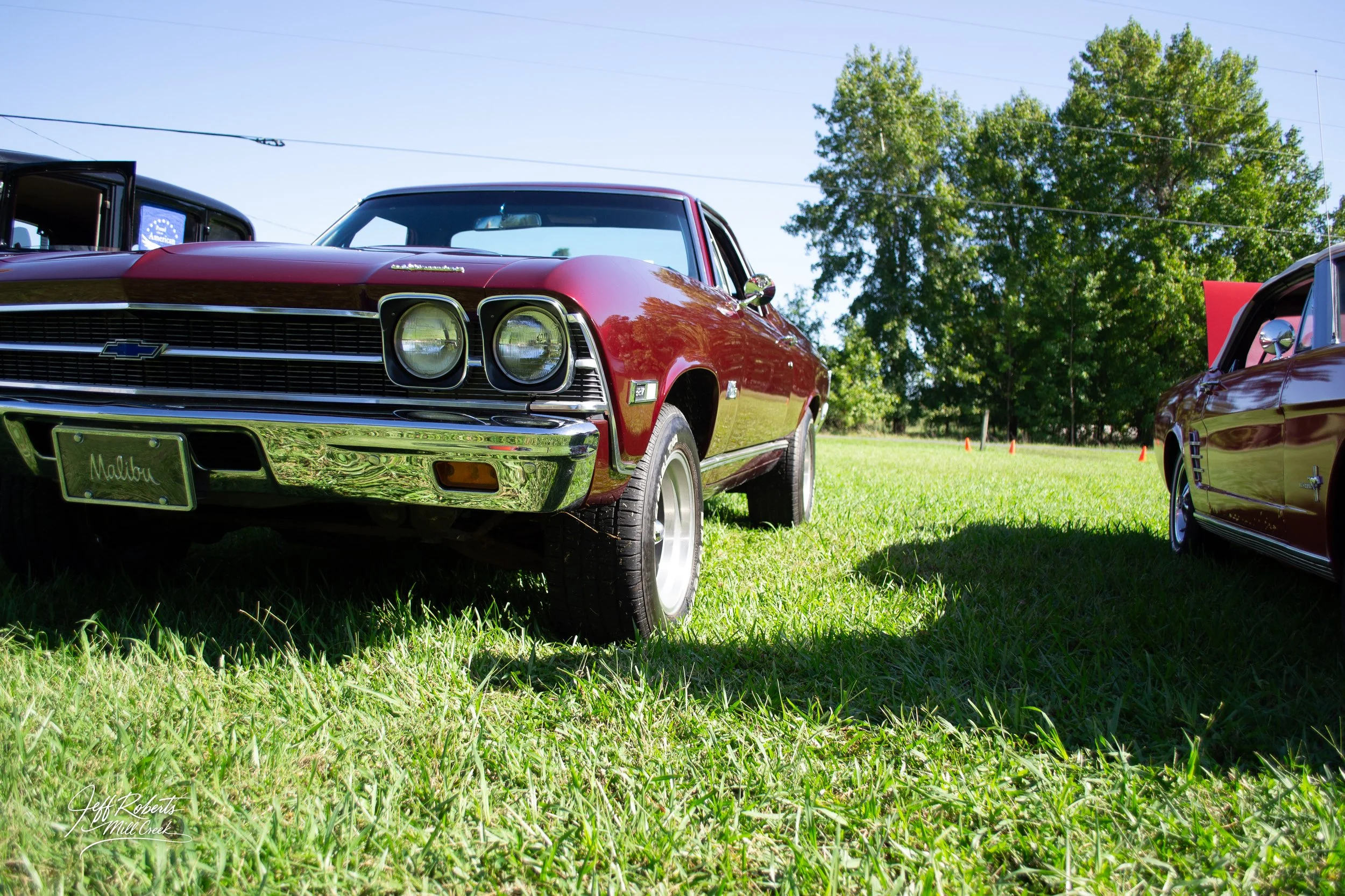 Red vintage Chevrolet Malibu car on grass field at a car show with trees in the background.