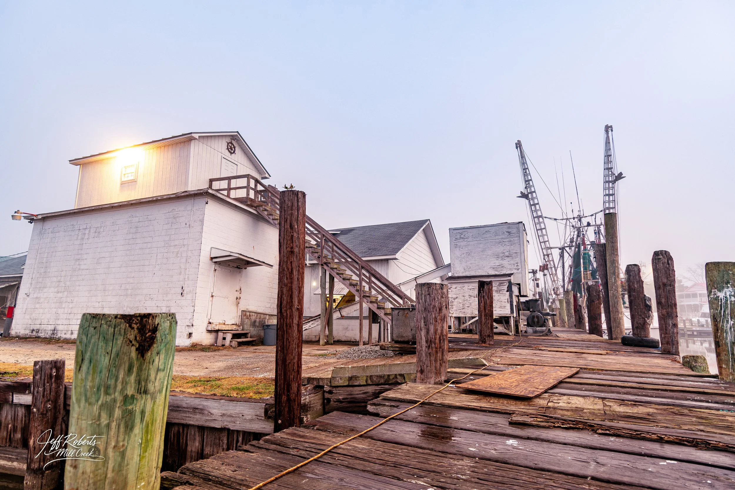 A weathered wooden dock with vertical posts, leading to a white building on stilts with a staircase, on a foggy day. In the background, there are fishing boats with tall masts and rigging.