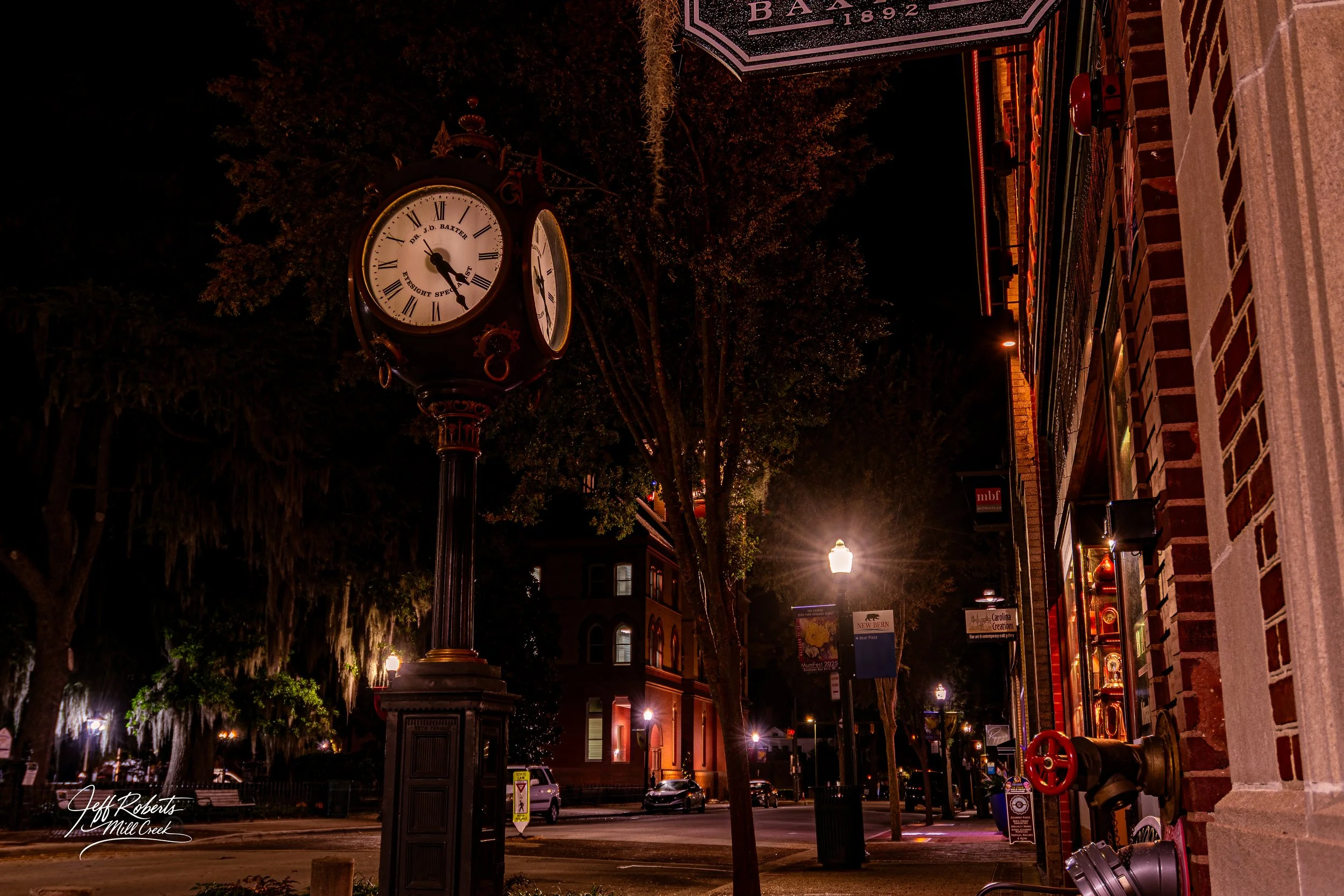 Night street scene with vintage clock on post, illuminated storefronts, trees, streetlights, and parked cars.