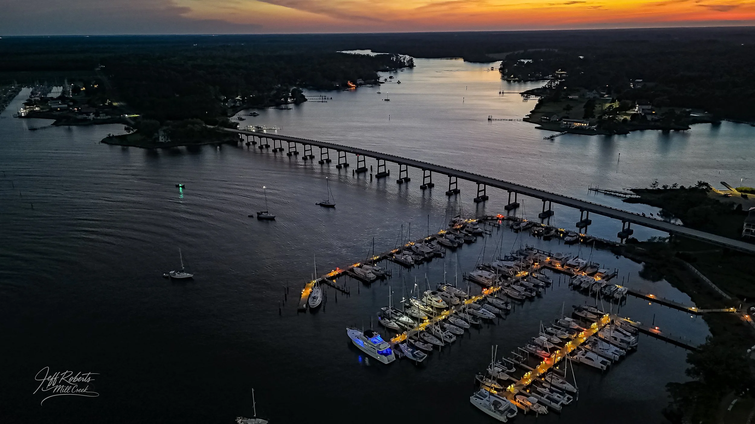 Aerial view of a marina with boats docked at night, illuminated by yellow lights. A bridge crosses the waterway in the background, with houses and trees lining the shoreline. The sky shows a sunset with orange and purple hues.