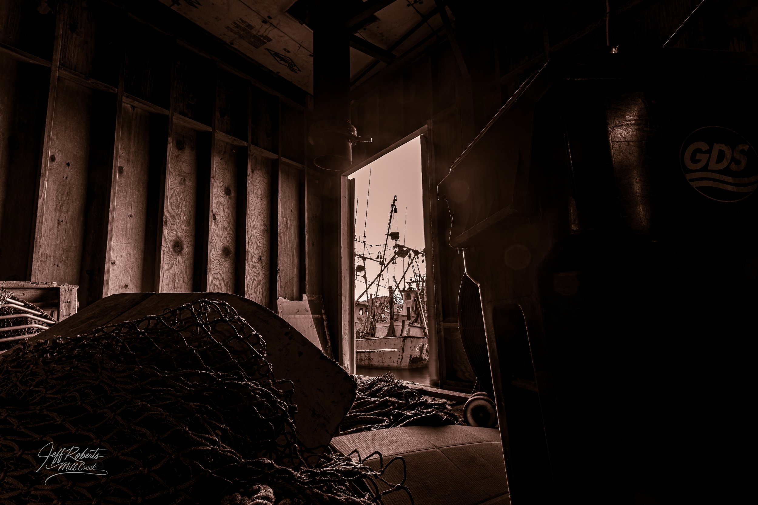 A dark boat shed with fishing gear and a boat visible outside, illuminated by natural light coming through an open door.