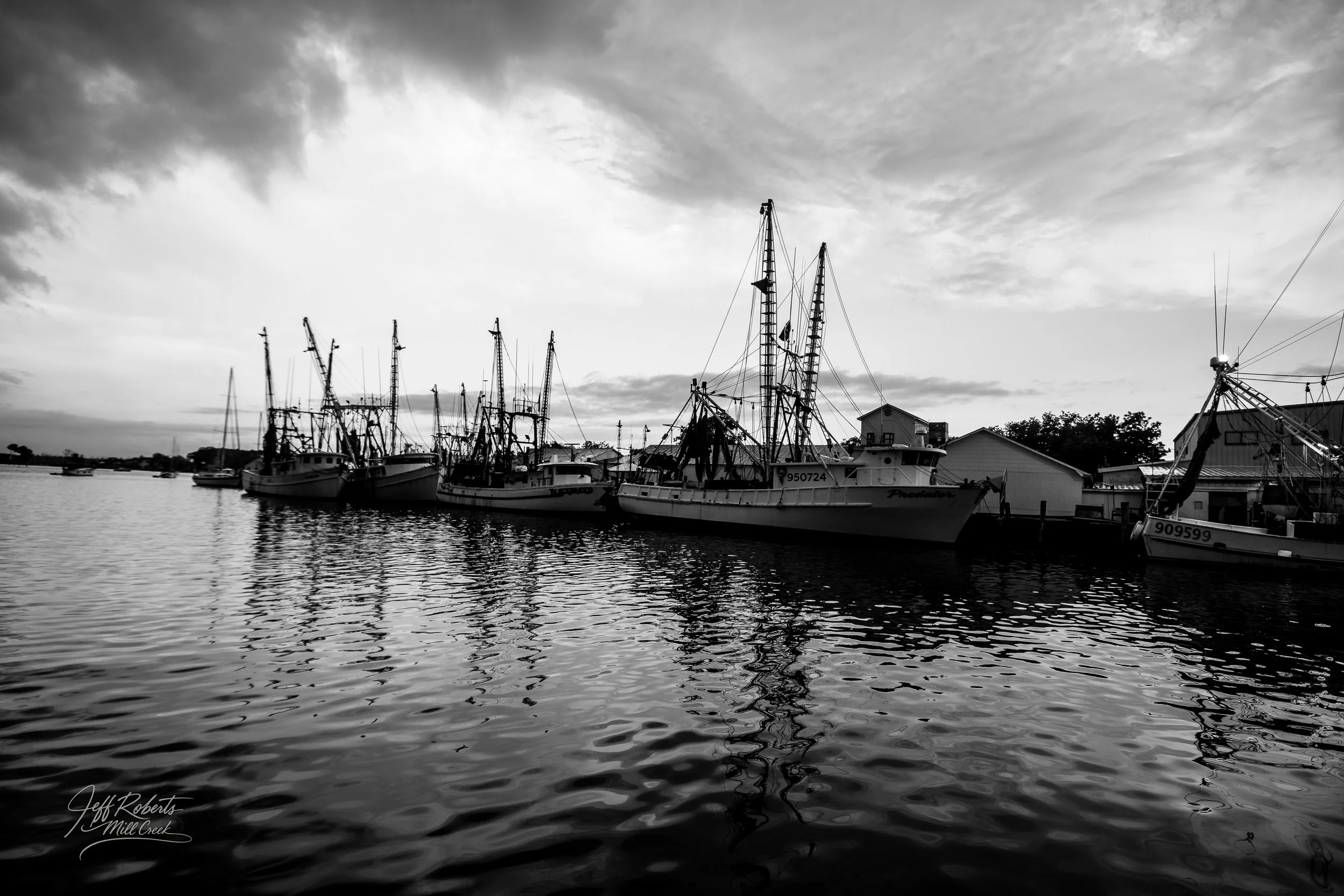 Black and white photo of several boats docked at a marina with calm water reflecting the boats, and a cloudy sky overhead.