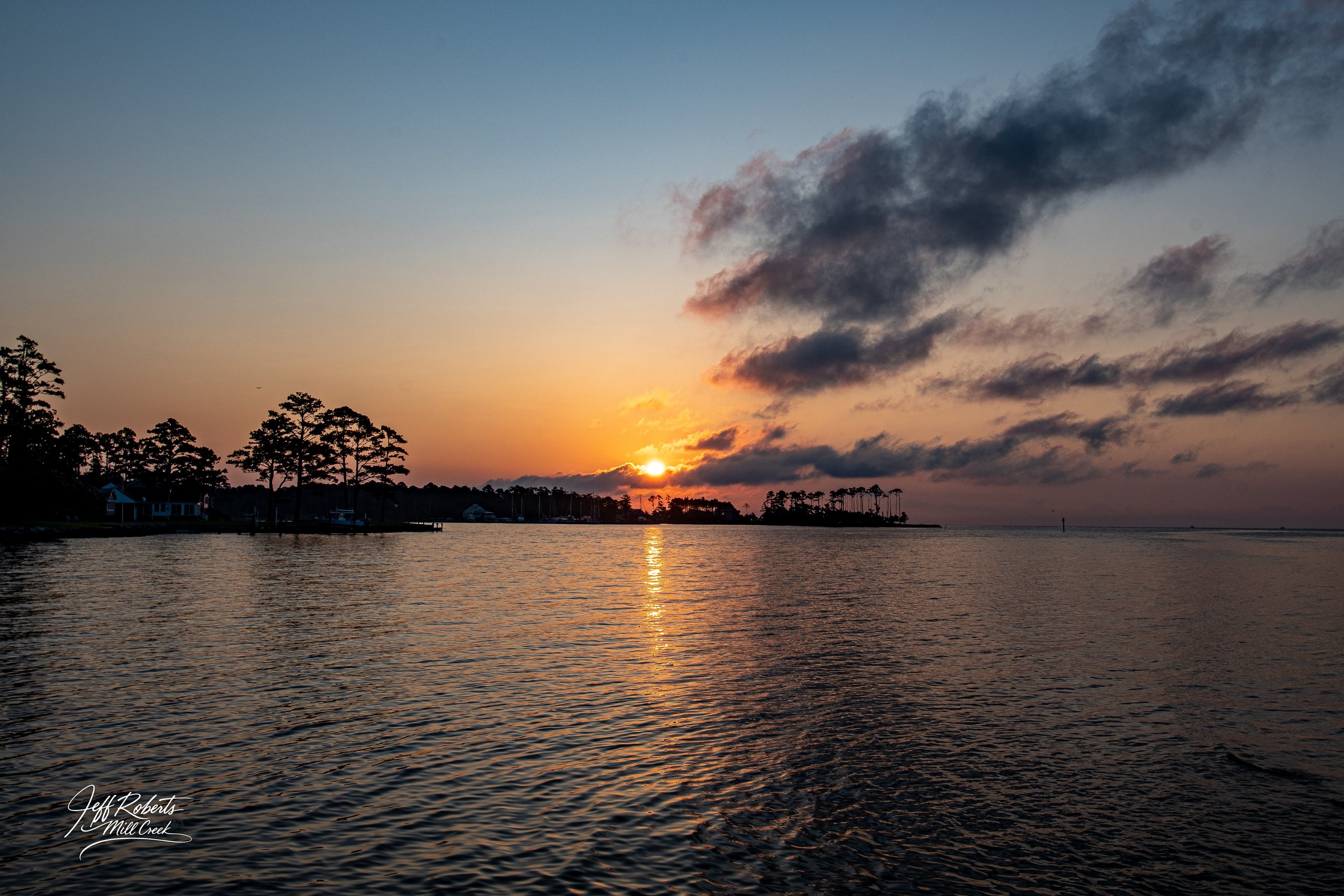 Sunset over a calm body of water with silhouettes of trees and a few houses along the shoreline, and dark clouds in the sky.